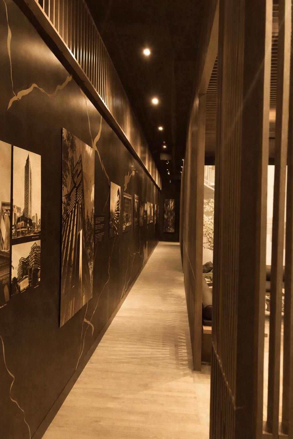 A narrow hallway with a black wall decorated with framed photographs of city buildings, illuminated by spotlights on the ceiling. On the right side, there is a wooden slat partition casting shadows on the light-colored wooden floor.