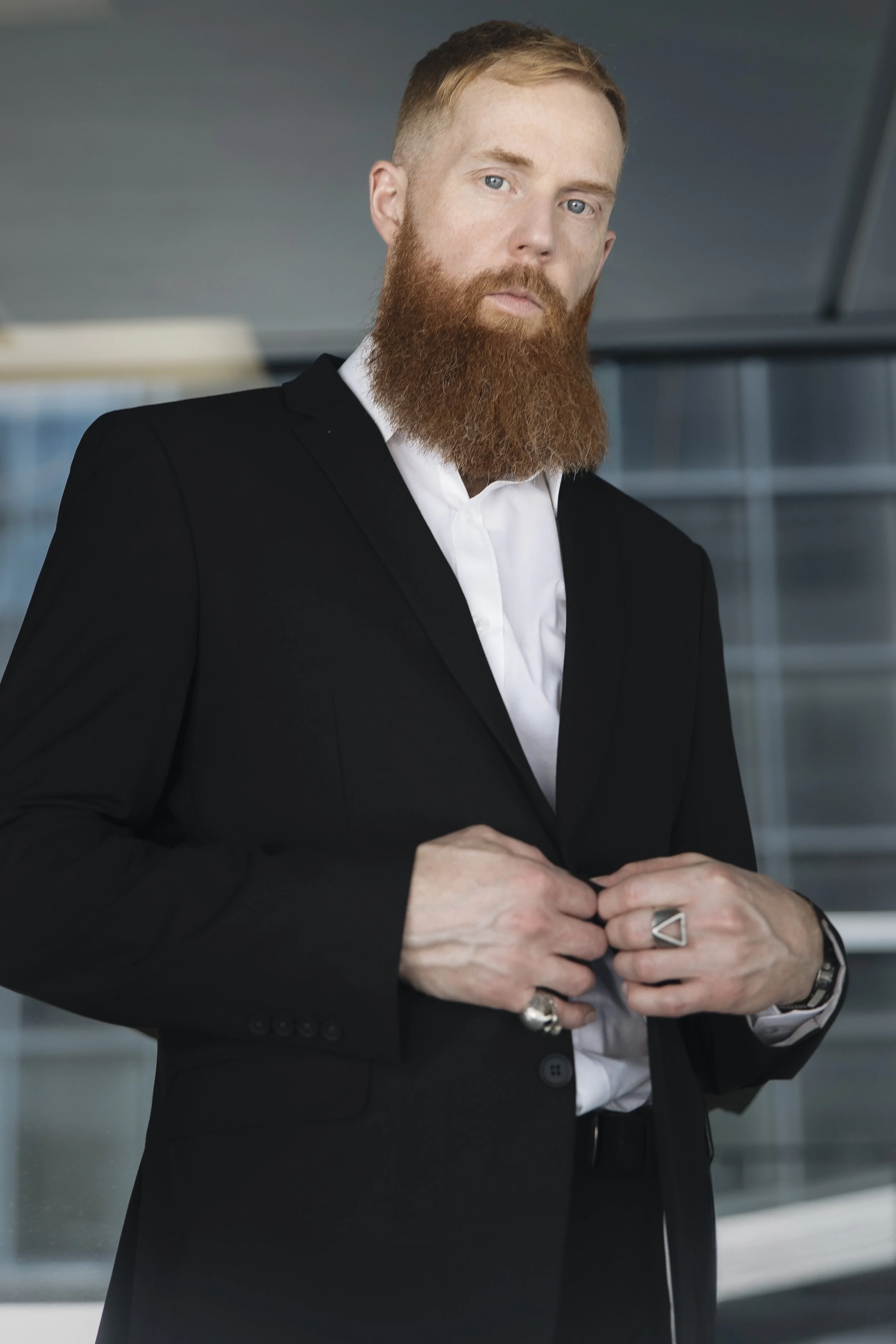 Man with red hair and beard wearing black suit and white shirt adjusting coat in a modern building.