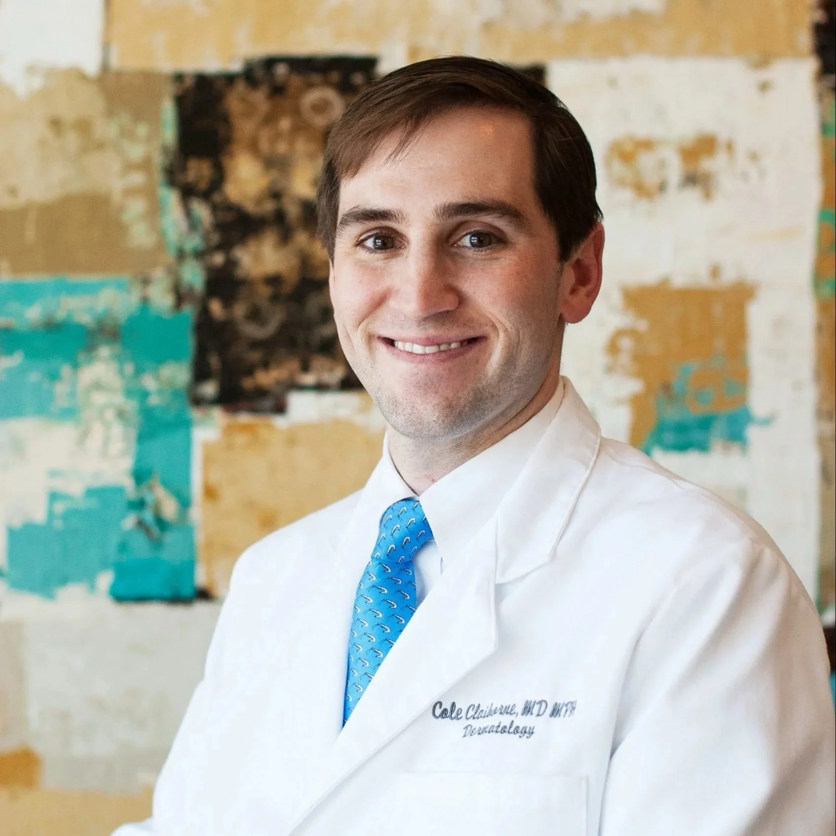 A smiling man in a white medical coat with blue tie, standing in front of an abstract multicolored artwork.
