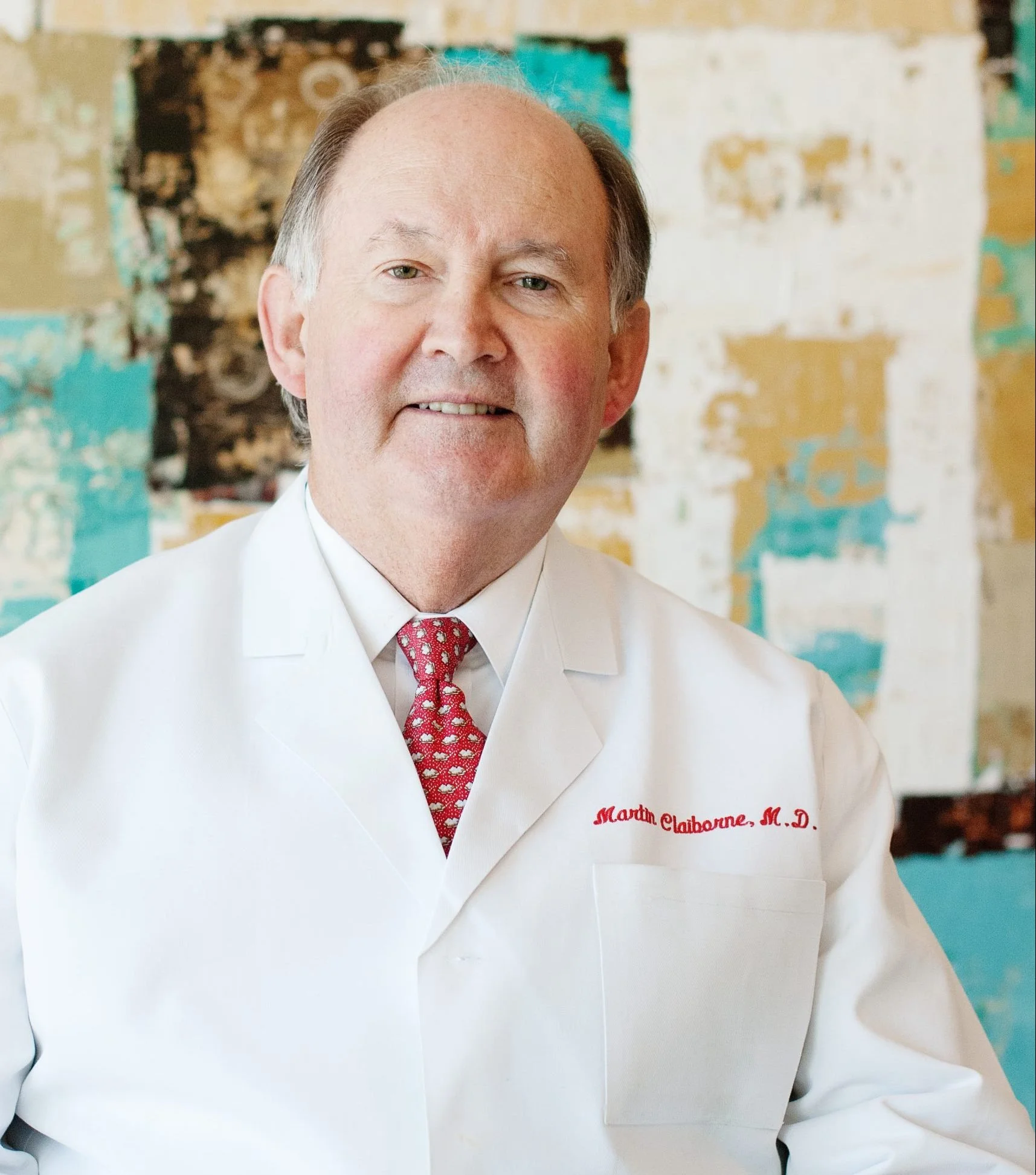 A senior man with gray hair, wearing a white lab coat with red embroidery, and a red patterned tie, posing in front of an abstract patterned background.