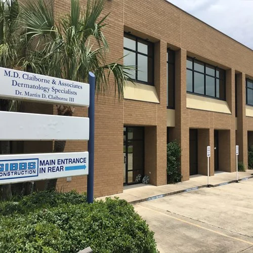 Building with signs for M.D. Claiborne & Associates dermatology practice and a construction company, with parking spaces and plants outside.
