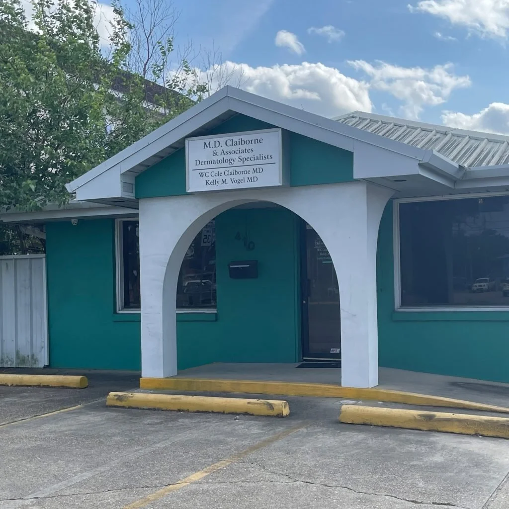Front view of a small teal building with white trim, housing dermatology specialists. The building has a sign with the names of the dermatology specialists, including M.D. Cole Claiborne and Kelly M. Vogel. There are parking spaces with yellow wheel stops in front of the building and a cloudy sky overhead.