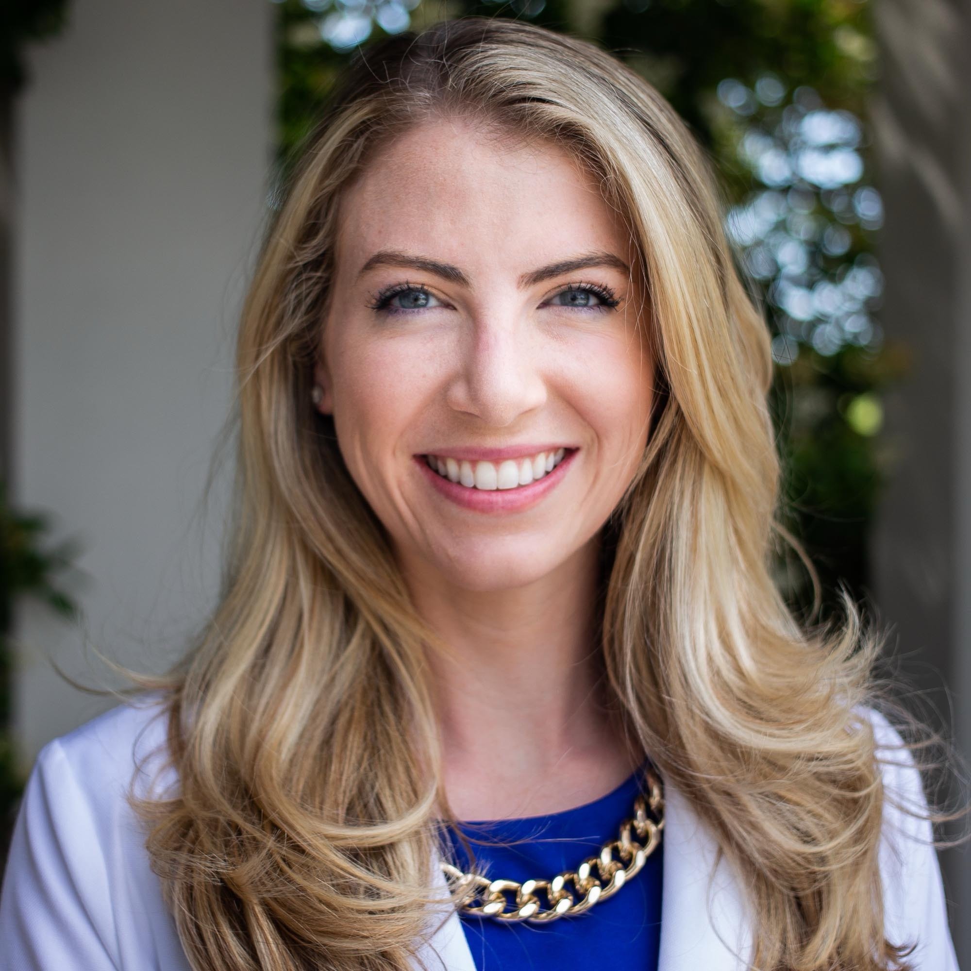 Close-up outdoor portrait of a young woman smiling with blonde hair, blue eyes, wearing a white blazer, blue top, and a gold chain necklace.