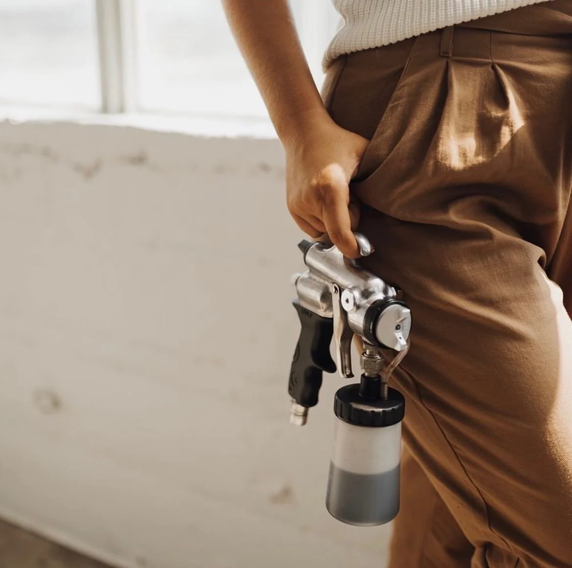 Image showing a female hand holding a spray tan gun by one finger with thumb hooked into a pair of tan brown pants.