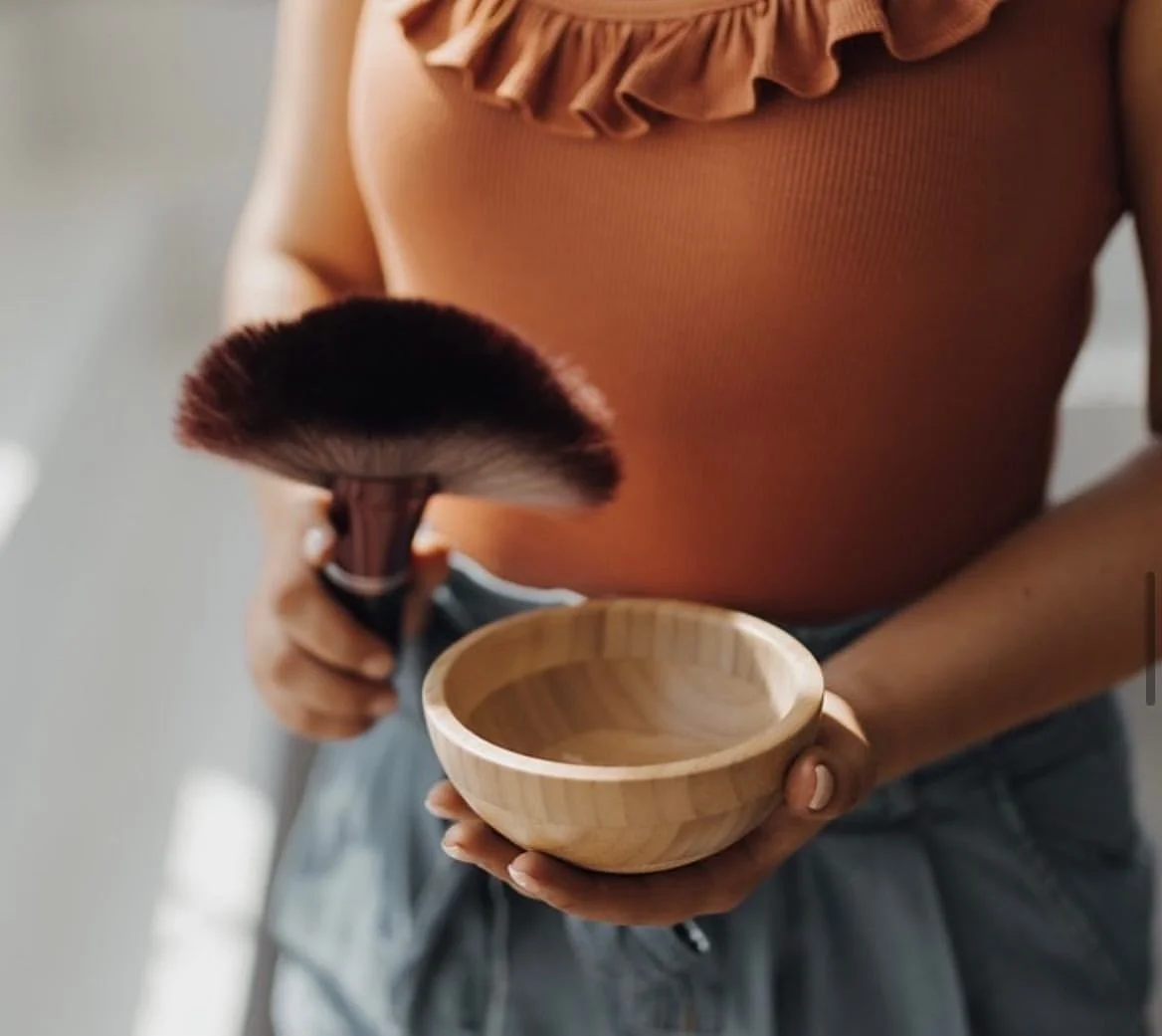 Female spray tan technician holding bowl of tanning powder and brush wearing ochre coloured top.