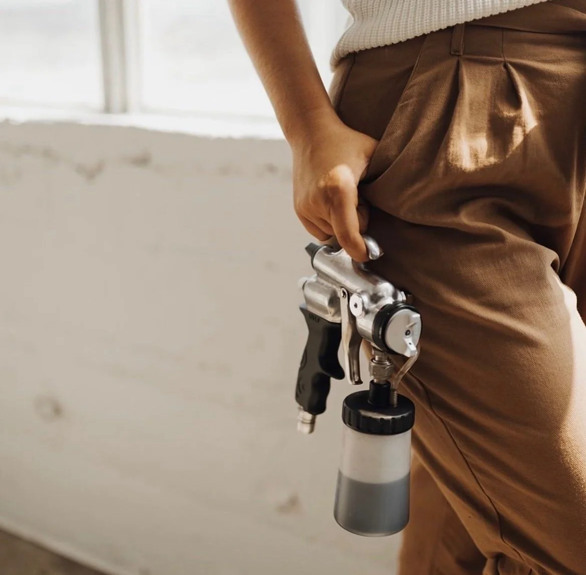 Image of spray tan technician's hand holding spray tanning gun wearing tan pants.