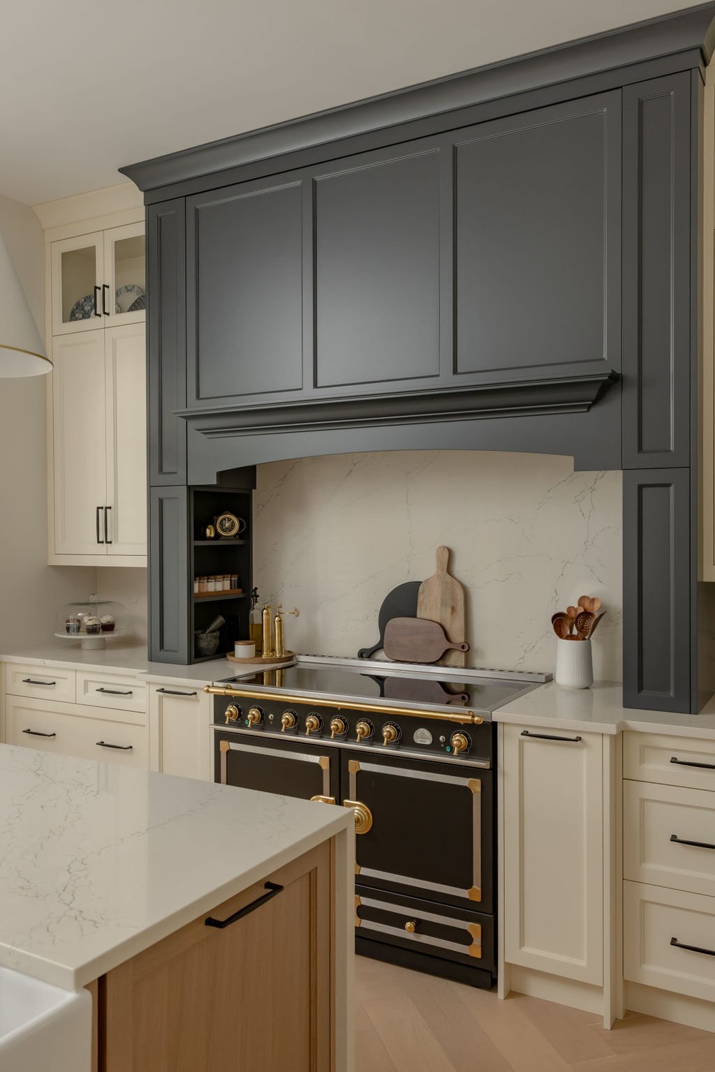 Modern kitchen with cream lower cabinets, a black and gold stove, and dark gray upper cabinetry. Featuring cornufe 110 induction range in black with gold and chrome hardware Decorative cutting boards and utensils are on the countertop.