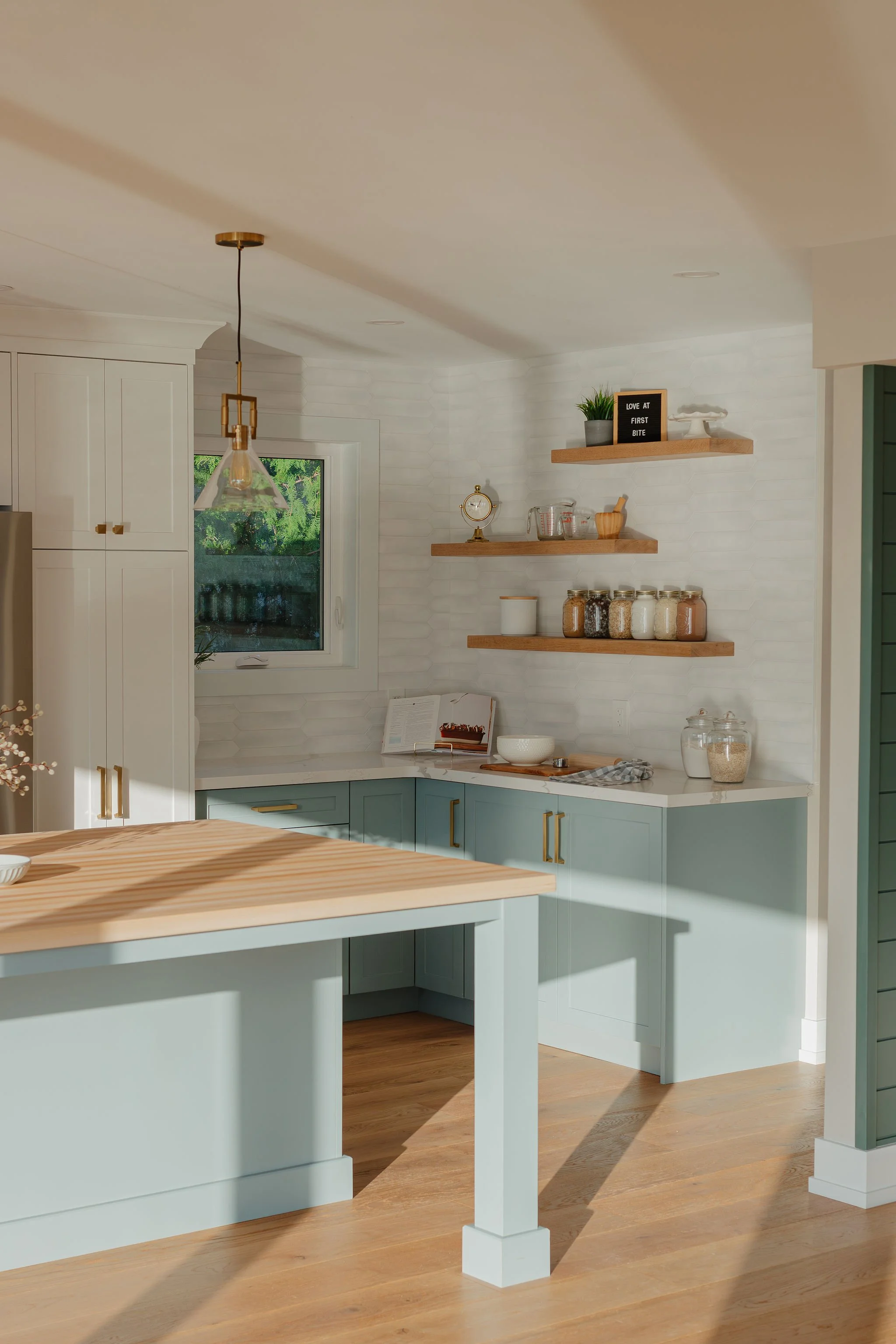 A bright kitchen with white cabinets and light teal lower cabinets, wooden open shelves holding jars and decorative items, a small window with greenery outside, and a white quartz countertop with a cookbook and bowl, illuminated by sunlight.