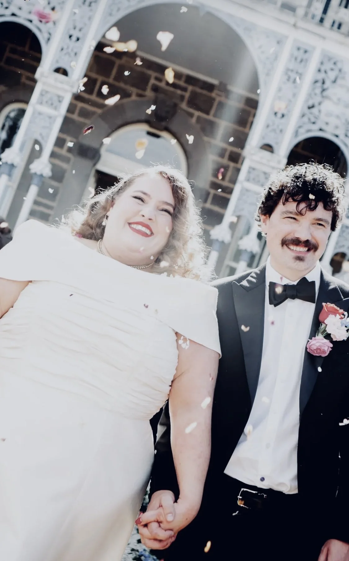 Happy couple in wedding attire holding hands outside, with confetti falling around them, in front of an ornate building entrance.