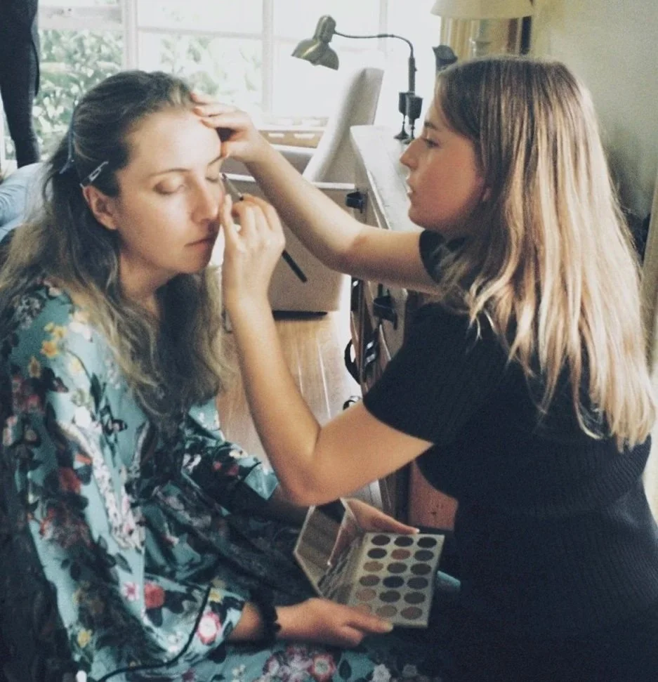 A woman sitting with her eyes closed as another woman in black applies makeup to her face, holding a makeup palette in her hand, inside a bright room with large windows.