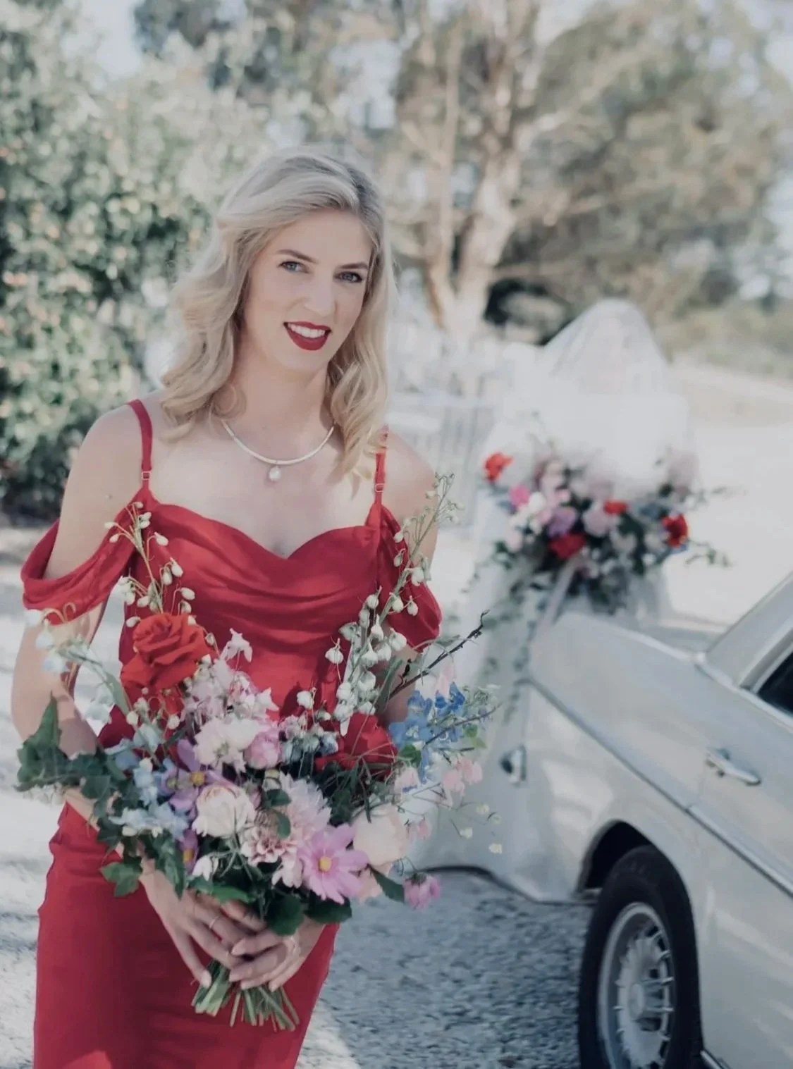 A woman with blonde hair wearing a red dress and holding a bouquet of colorful flowers, outdoors near a vintage white car decorated with flowers.