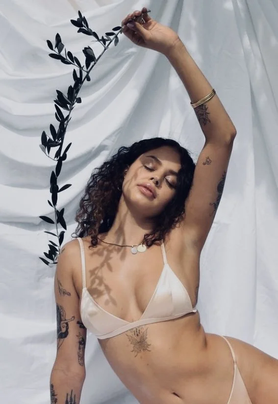 Woman with curly hair and tattoos, wearing lingerie, poses with one arm raised holding a branch with black leaves, against a white background.