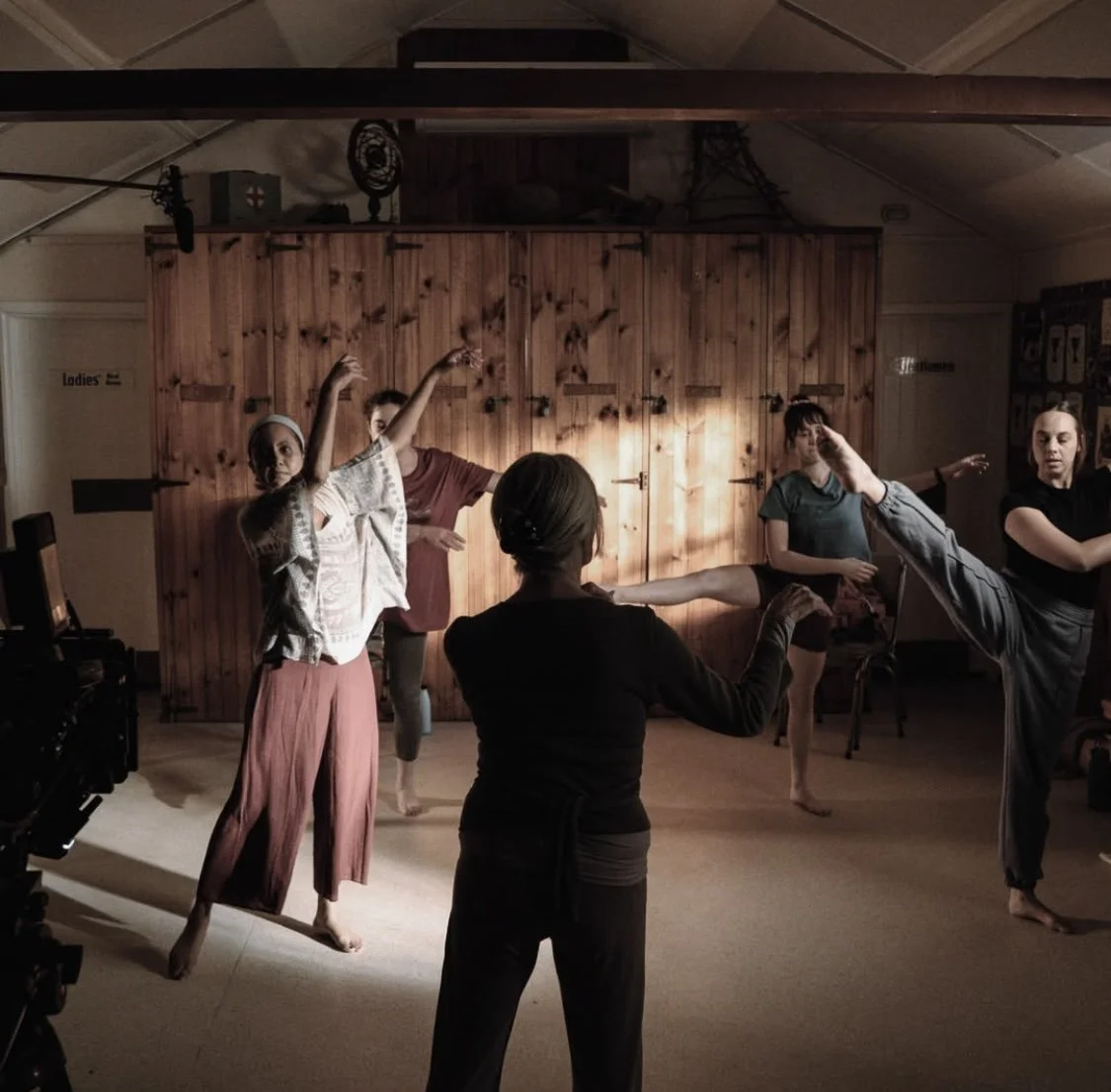 People practicing yoga indoors, with a wooden wall background, some standing on one leg in balancing poses.