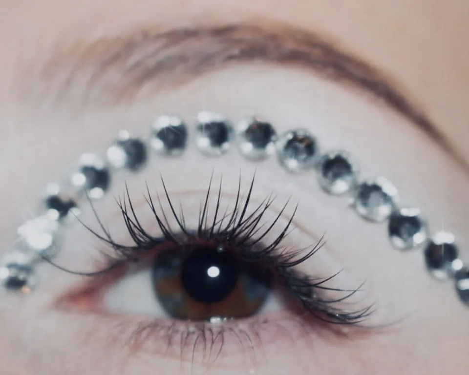 Close-up of an eye with long eyelashes, surrounded by a semi-circular arrangement of rhinestones or crystals.