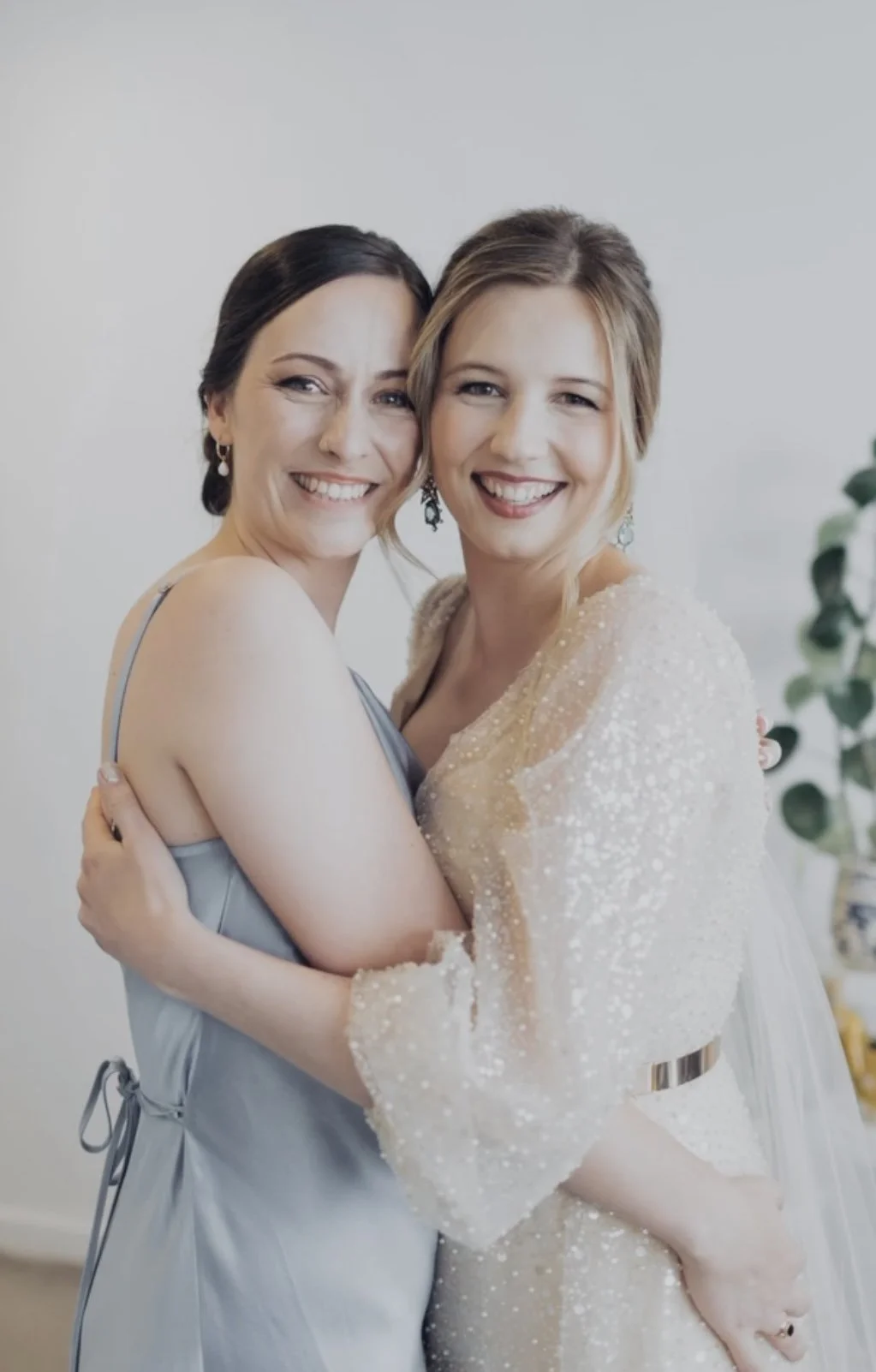 Two women smiling and hugging each other, one wearing a light blue dress and the other wearing a white dress with beaded details.