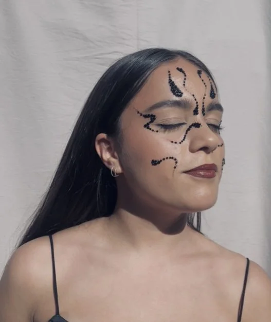 Woman with face makeup resembling a butterfly or face paint design, closed eyes, dark hair, earrings, and a black spaghetti strap top, standing in front of a plain white background.