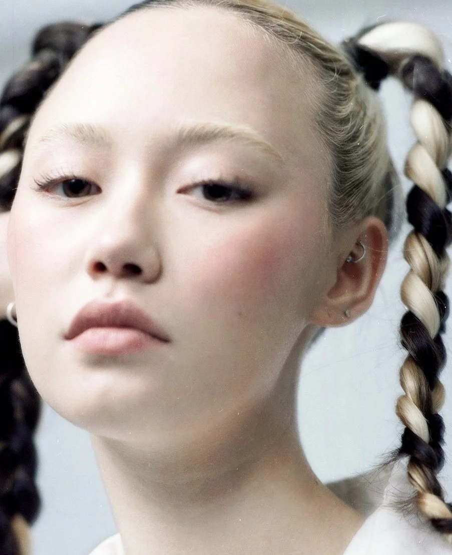 Close-up of a young woman with braided hair, wearing small earrings, and making a neutral facial expression.