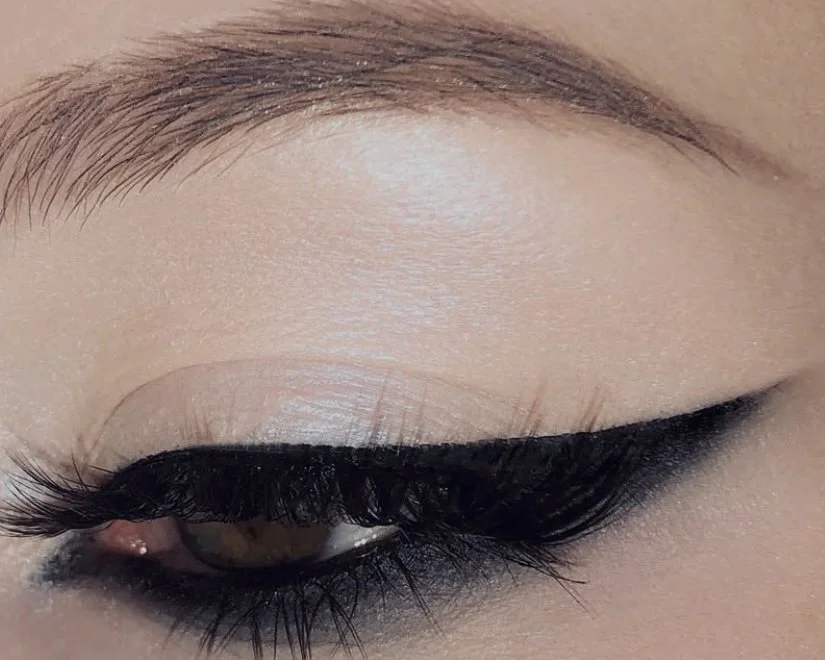 Close-up of a person's eye with black eyeliner and mascara, showing well-groomed eyebrows and long eyelashes.