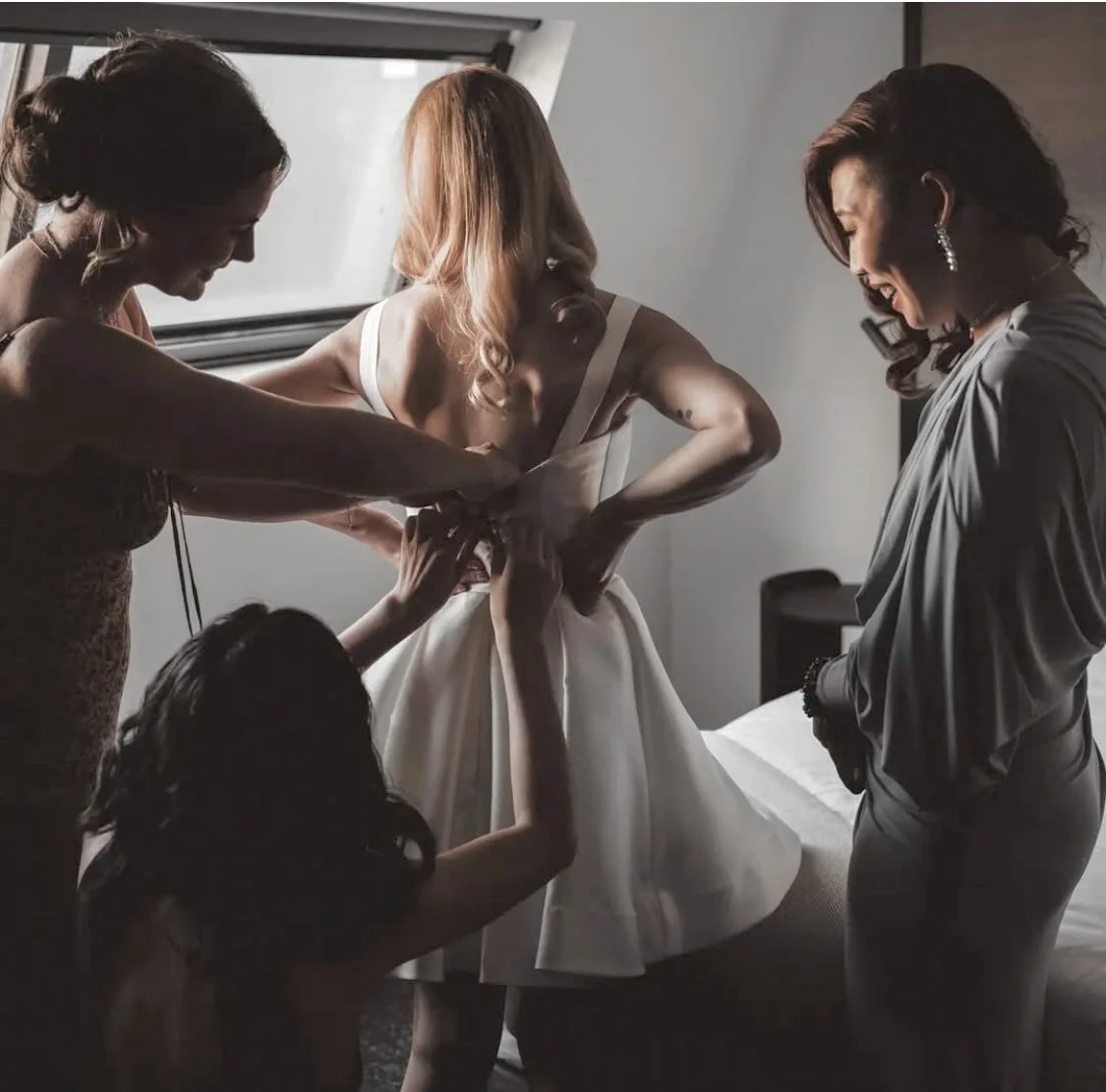 A bride wearing a white dress is being assisted by three women as they help her into her wedding gown in a softly lit room.