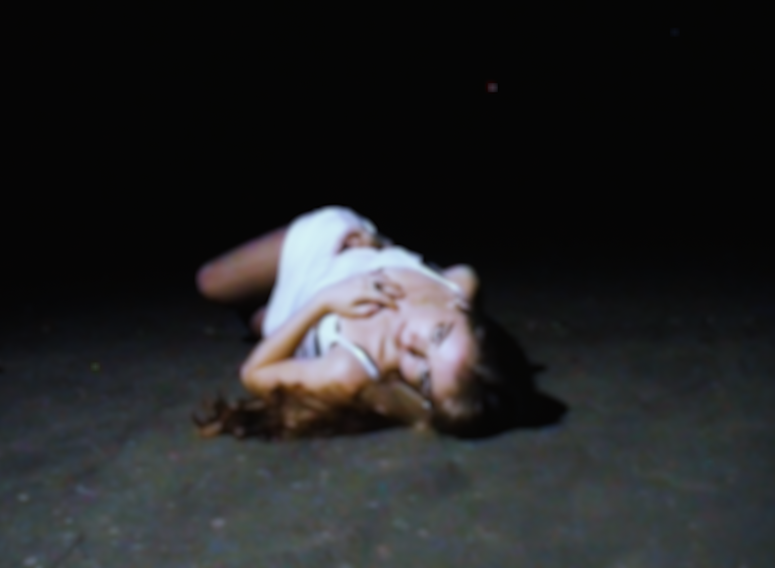 A woman lying on her side on a dark floor, her head resting on her arm, with blurred surroundings.