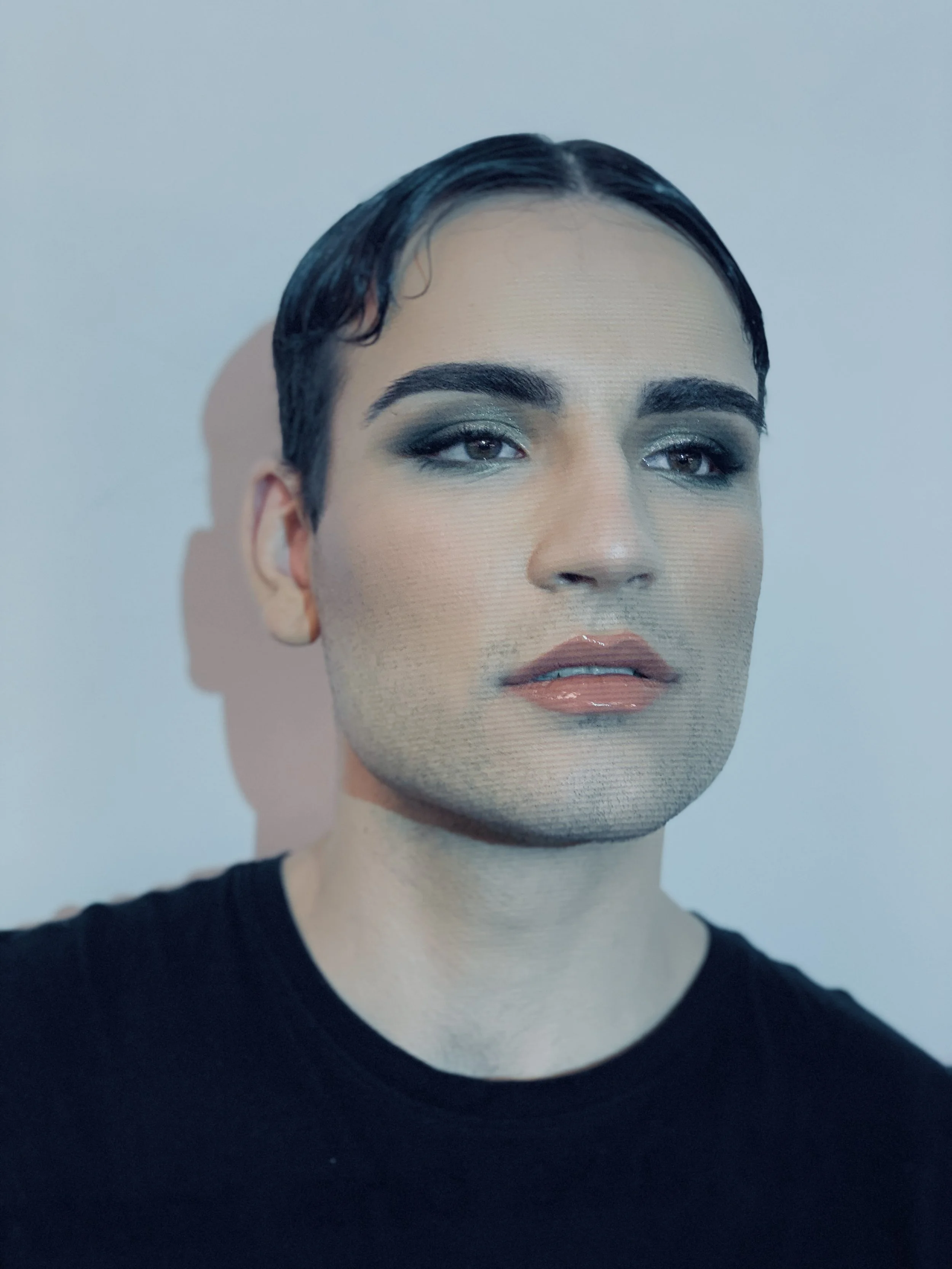 Close-up portrait of a person with short dark hair, bold eyebrows, blue and green eye makeup, glossy lips, and wearing a black shirt, standing against a light-colored background.