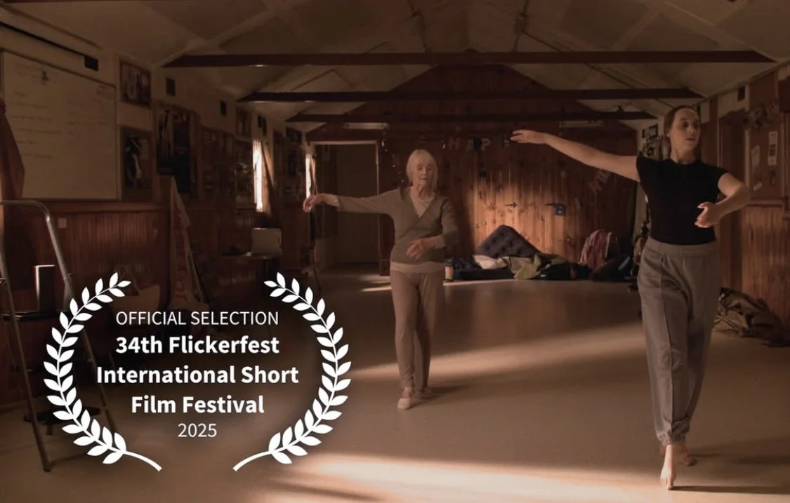 Two women dancing alone in a wooden-floored, barn-like room with warm lighting, a pile of clothes and a chair in the background, and a whiteboard on the wall. Text overlay states 'OFFICIAL SELECTION 34th Flickerfest International Short Film Festival 
