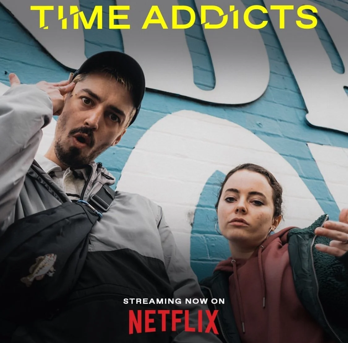 Two young adults, a man and a woman, taking a selfie in front of a blue wall with large white and blue text. The man is wearing a black cap and a gray jacket, and has a surprised expression. The woman is wearing a green jacket with a reddish hoodie a