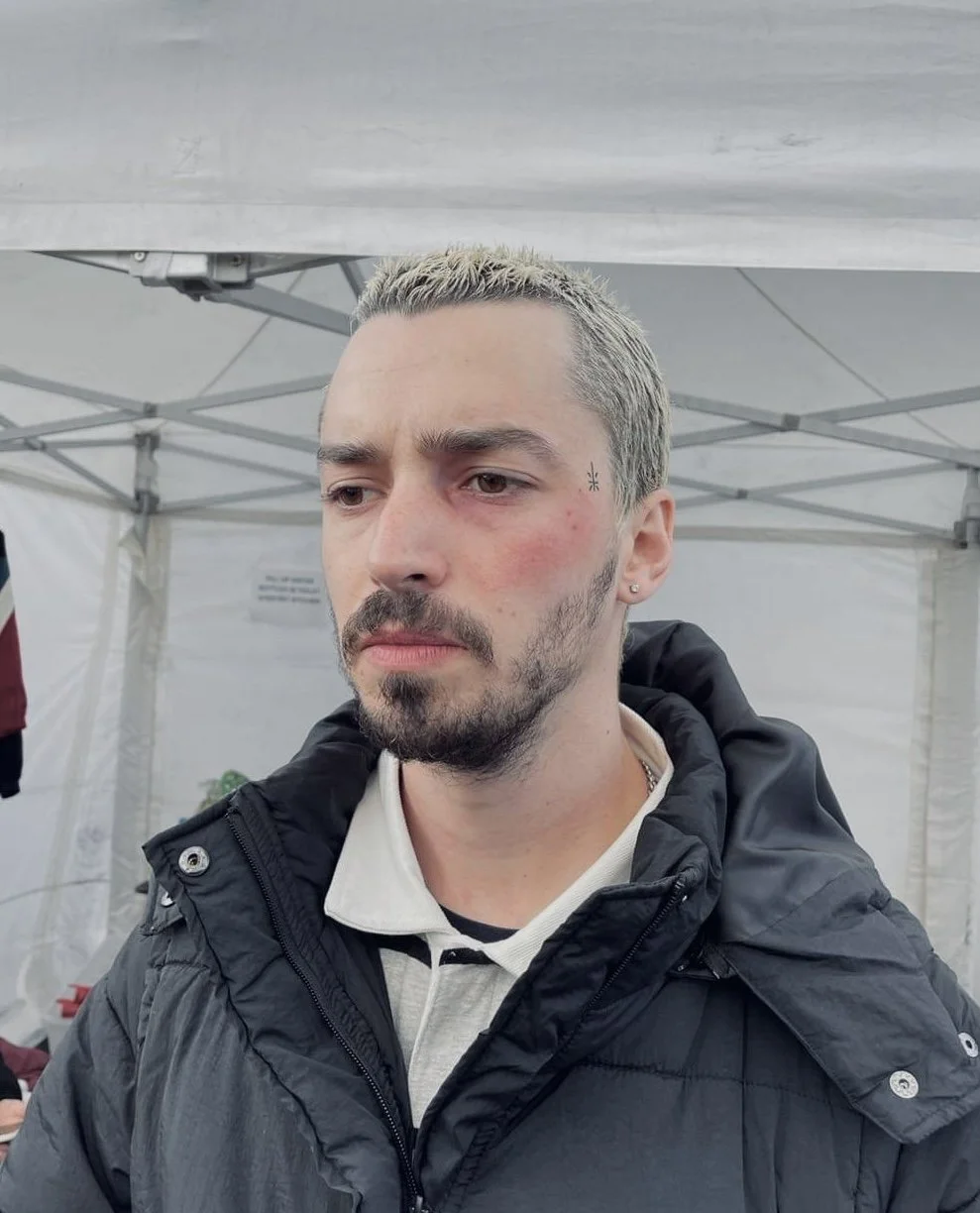 A man with short, light-colored hair, a beard, and mustache, wearing a black jacket over a beige polo shirt, standing under a white canopy tent.