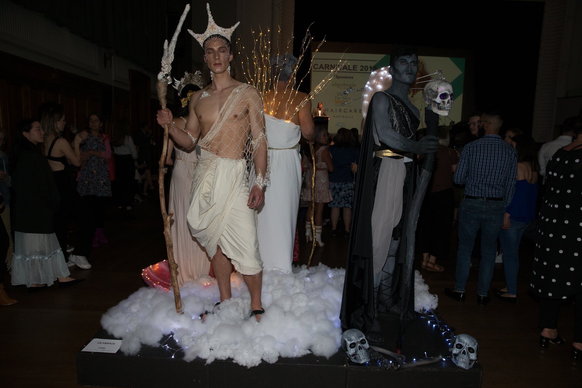 A display at an event featuring a model dressed as a mythological figure with a staff, standing on a cloud-like platform, next to a figure painted black with a skull mask, holding a scepter with a skull on top, surrounded by small skull decorations.