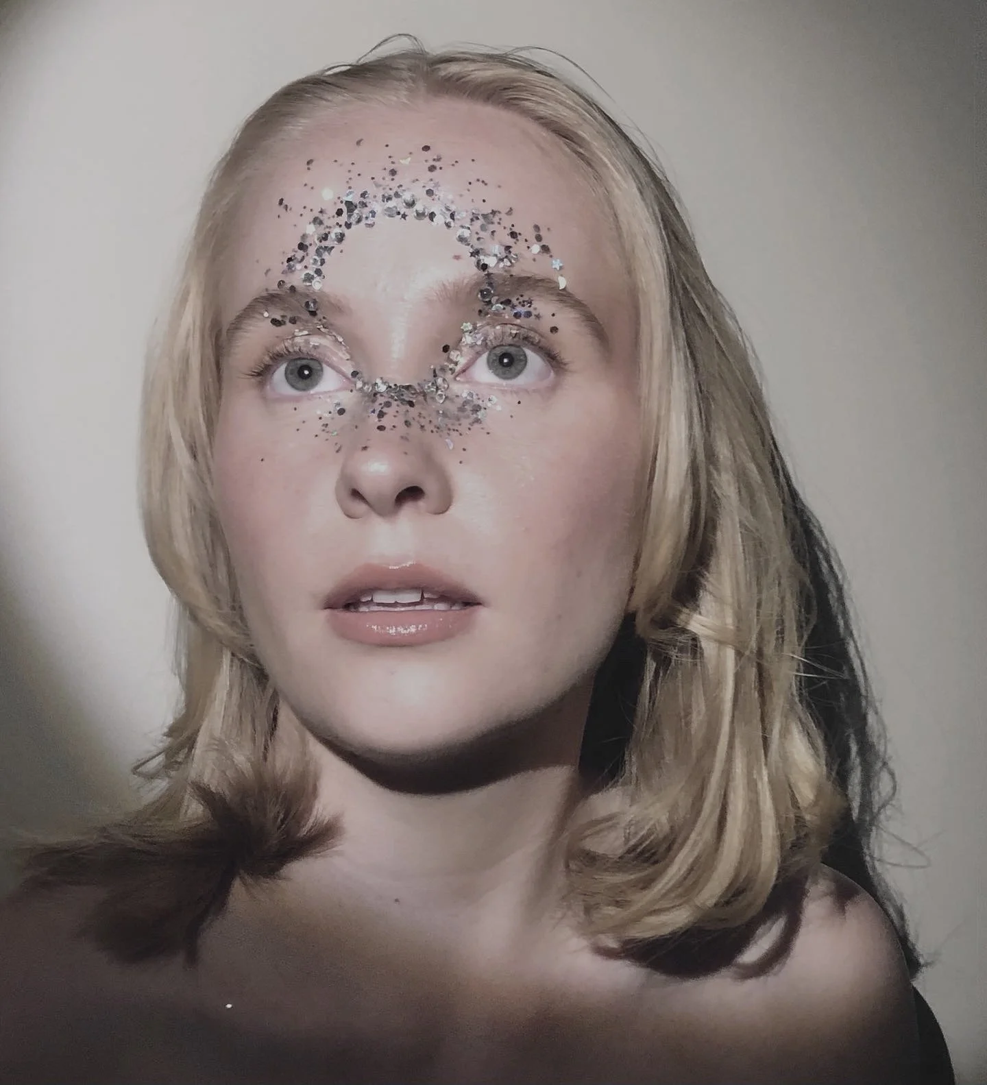 Close-up of a young woman with blonde hair and blue eyes, wearing face glitter arranged in a circular pattern around her eyes and nose, against a plain background.