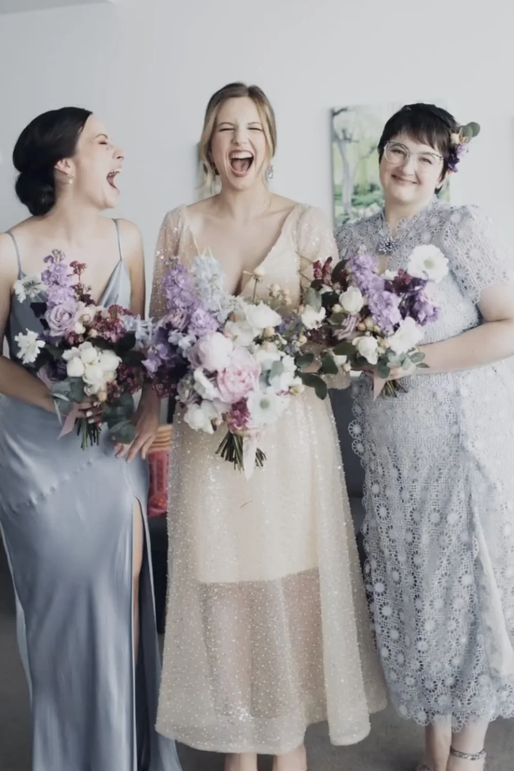 Three women in elegant dresses smiling, holding bouquets of purple and white flowers, during a celebration.
