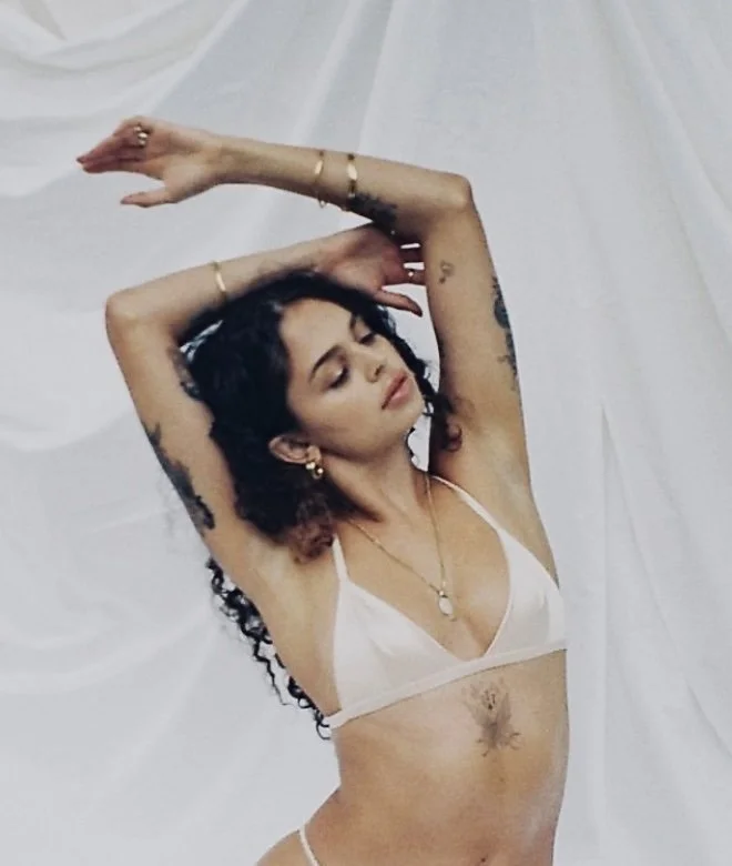 A woman with curly dark hair and tattoos poses with her arms over her head against a white fabric background, wearing a white bra and jewelry.