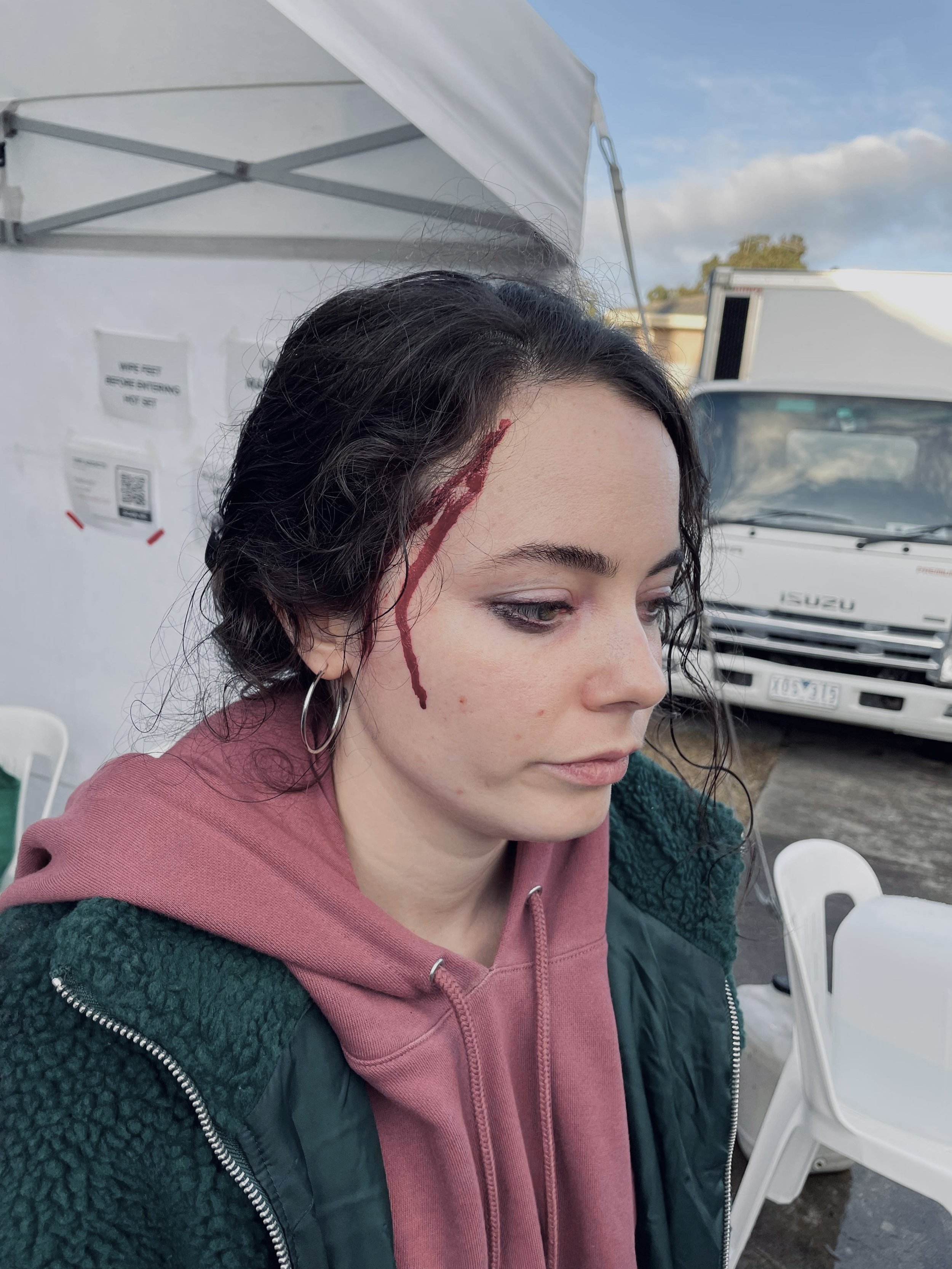 A young woman with dark curly hair and hoop earrings has fake blood running down the side of her face near her forehead, looking downwards with a neutral expression. She is wearing a pink hoodie and a dark green jacket, with a white tent and trucks i