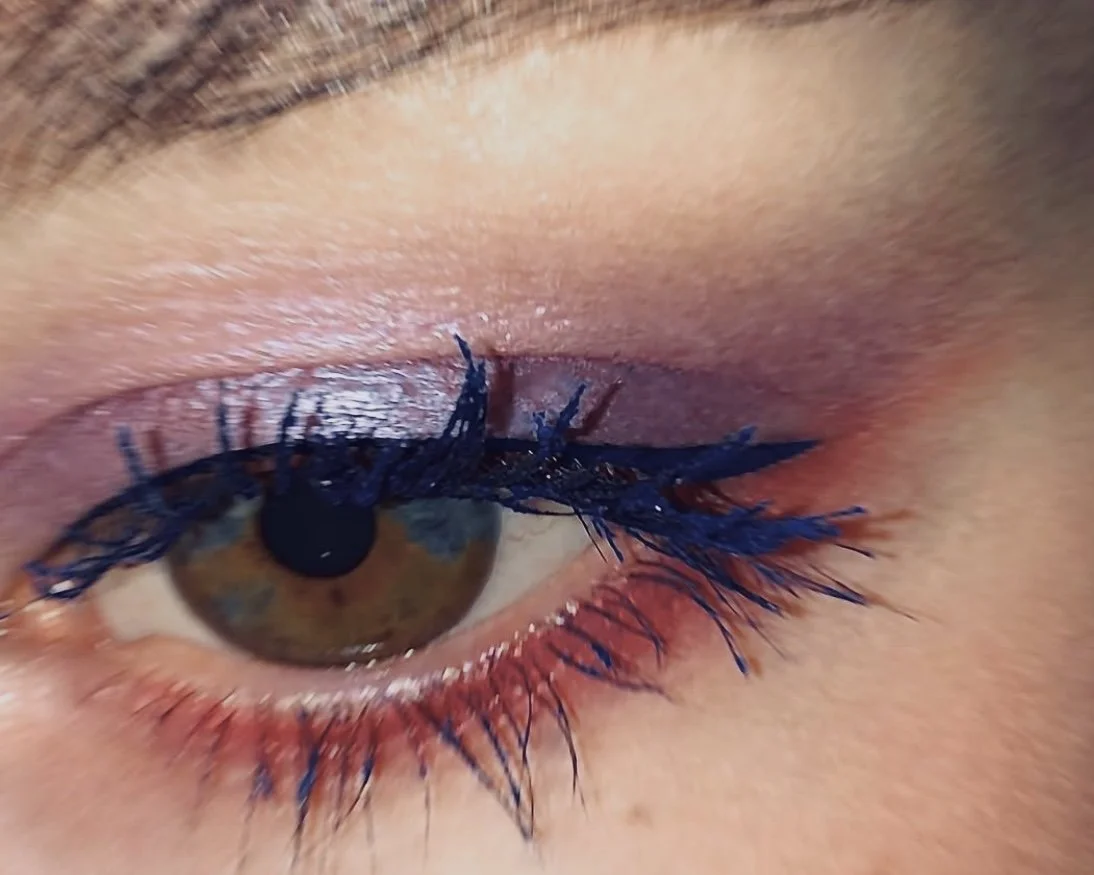 A close-up of a person's eye with blue feathered false eyelashes and makeup, including pink eyeshadow.