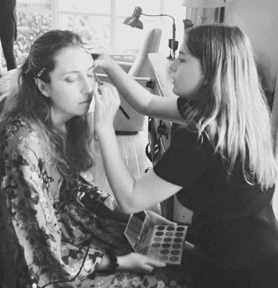 A woman is applying makeup to another woman sitting with her eyes closed. The makeup artist is holding a palette of eyeshadows and is using a makeup brush, while the seated woman appears to be getting ready for an event.