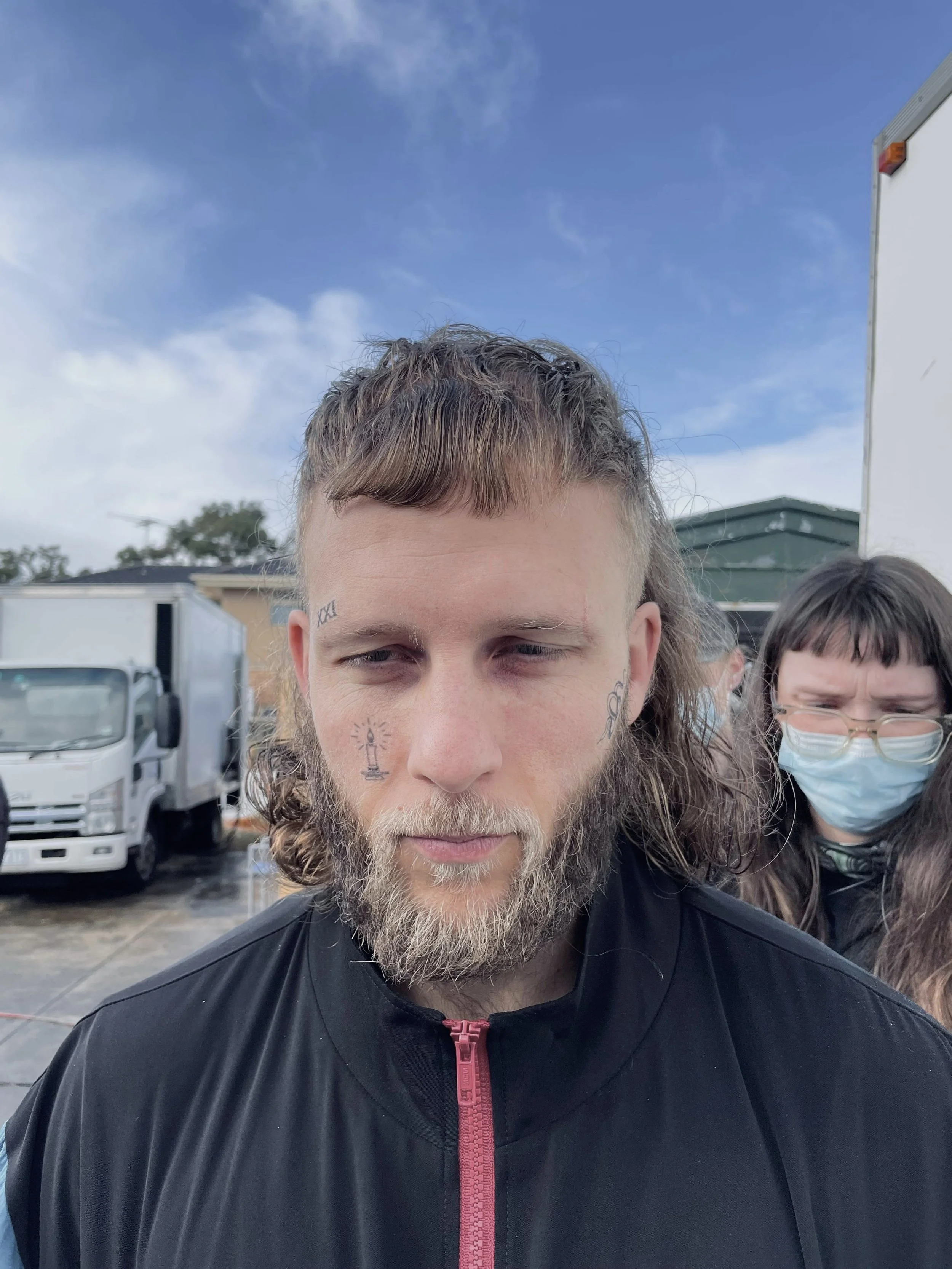 A young man with a beard and tattoos on his face, wearing a black jacket with a pink zipper, stands outside on a cloudy day. There are trucks and other people, some wearing masks, in the background.