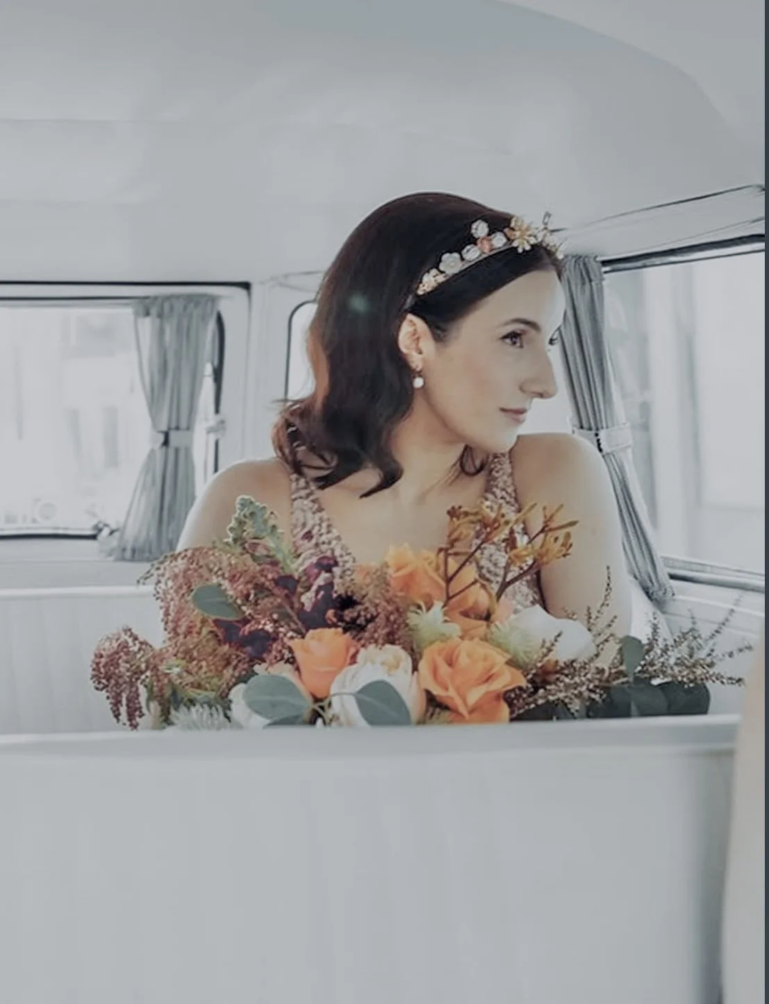 A woman with dark hair, light skin, wearing a floral headband and pearl earrings, sitting in a vehicle, holding a bouquet of orange, white, and green flowers.