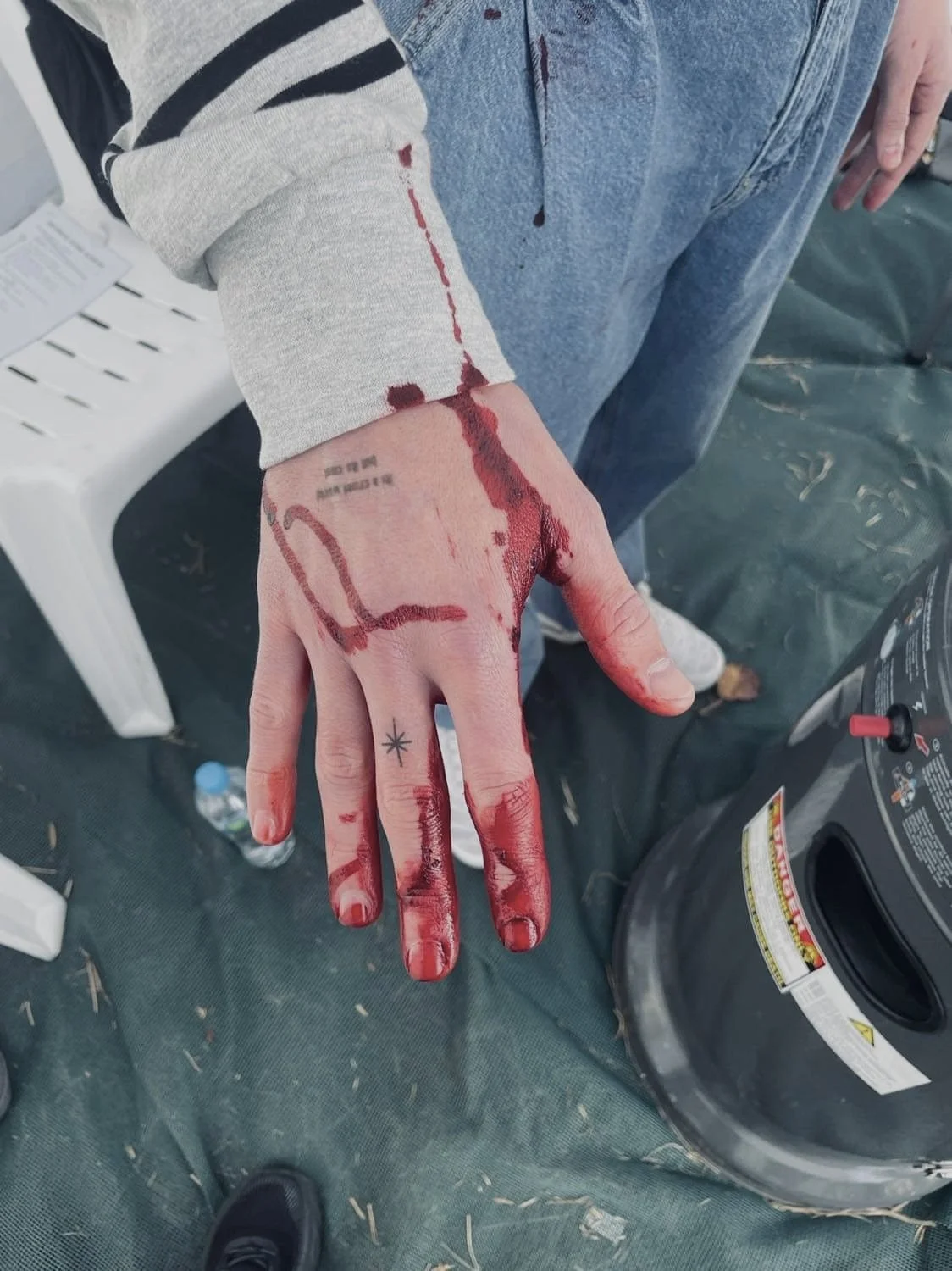 Person with blood on their hand and arm, showing bloodstains and a small tattoo of a star on their finger, standing near a plastic chair and a propane tank.