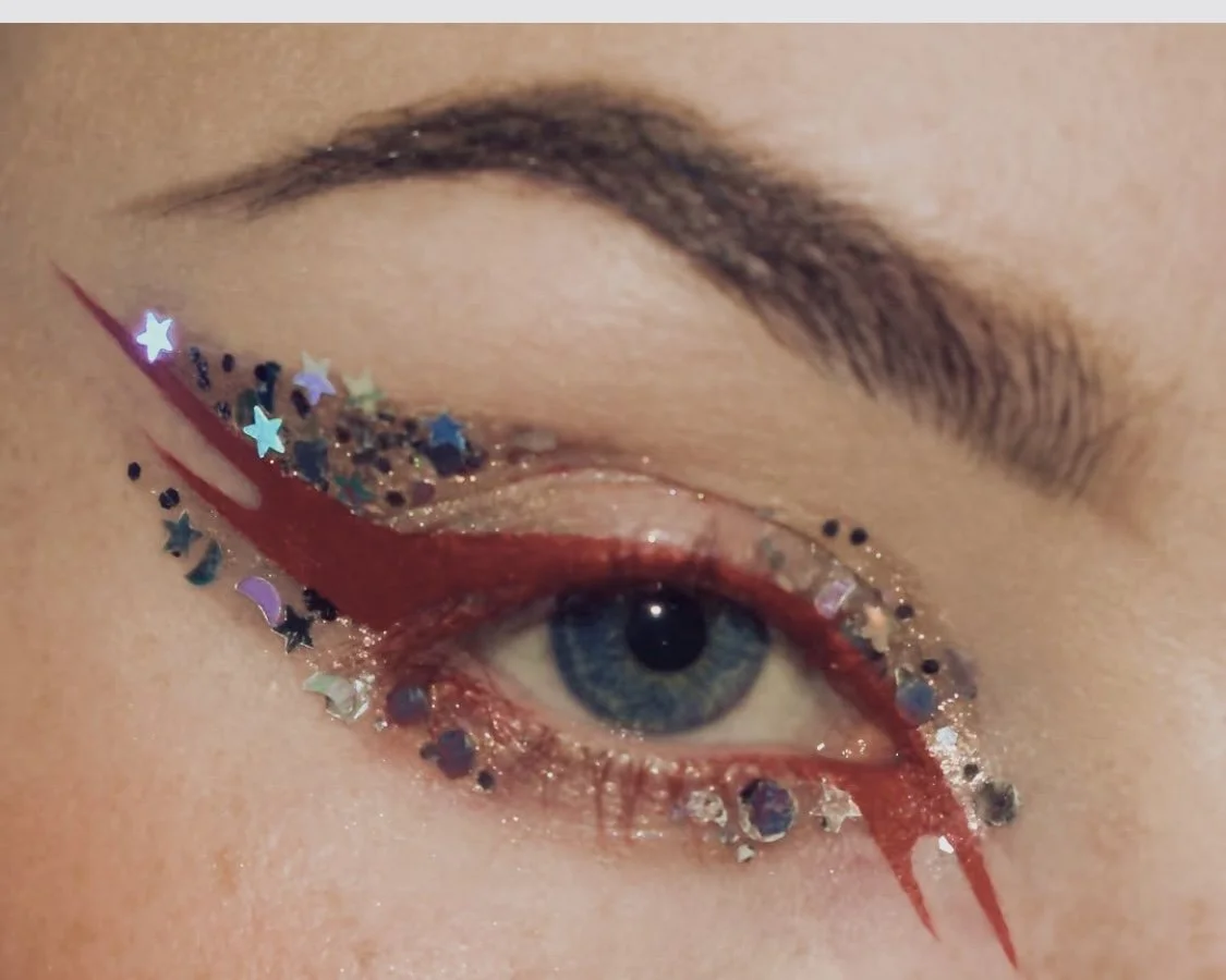 Close-up of a person’s eye with artistic makeup featuring glitter and star-shaped confetti, with a bold red line extending from the corner of the eye.