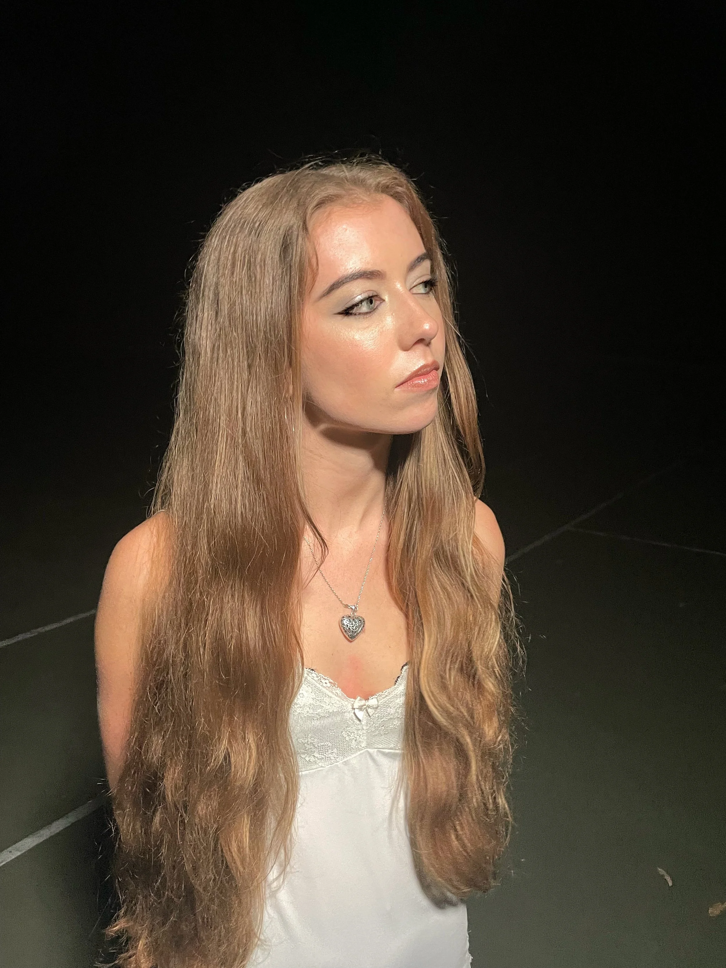 A young woman with long, wavy blonde hair wearing a white lace-trimmed top and a silver heart-shaped pendant necklace, standing against a black background.