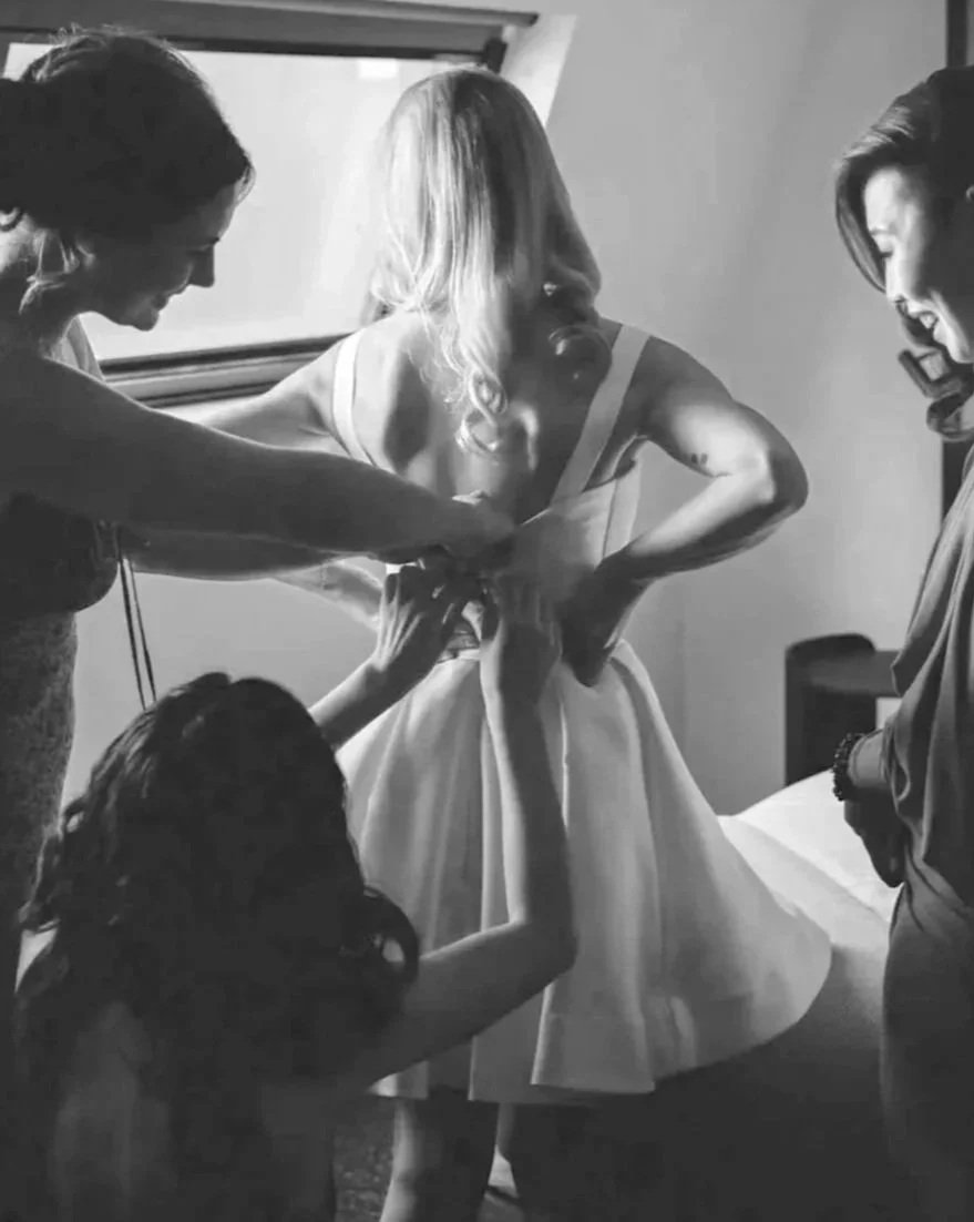 Women helping a bride put on her wedding dress, with a flower girl helping with the dress train, in a dressing room.
