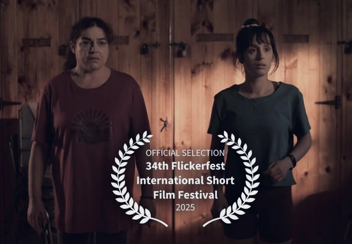 Two women standing in a wooden room, one appears older and the other younger, with the girl wearing a ponytail and casual clothing. An award message overlay indicates the film's selection at the 34th Flickerfest International Short Film Festival in 2