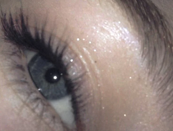 Close-up of a human eye, showing lashes, eyelid, and iris in shades of gray, with some sparkles or glitter near the eyelid.
