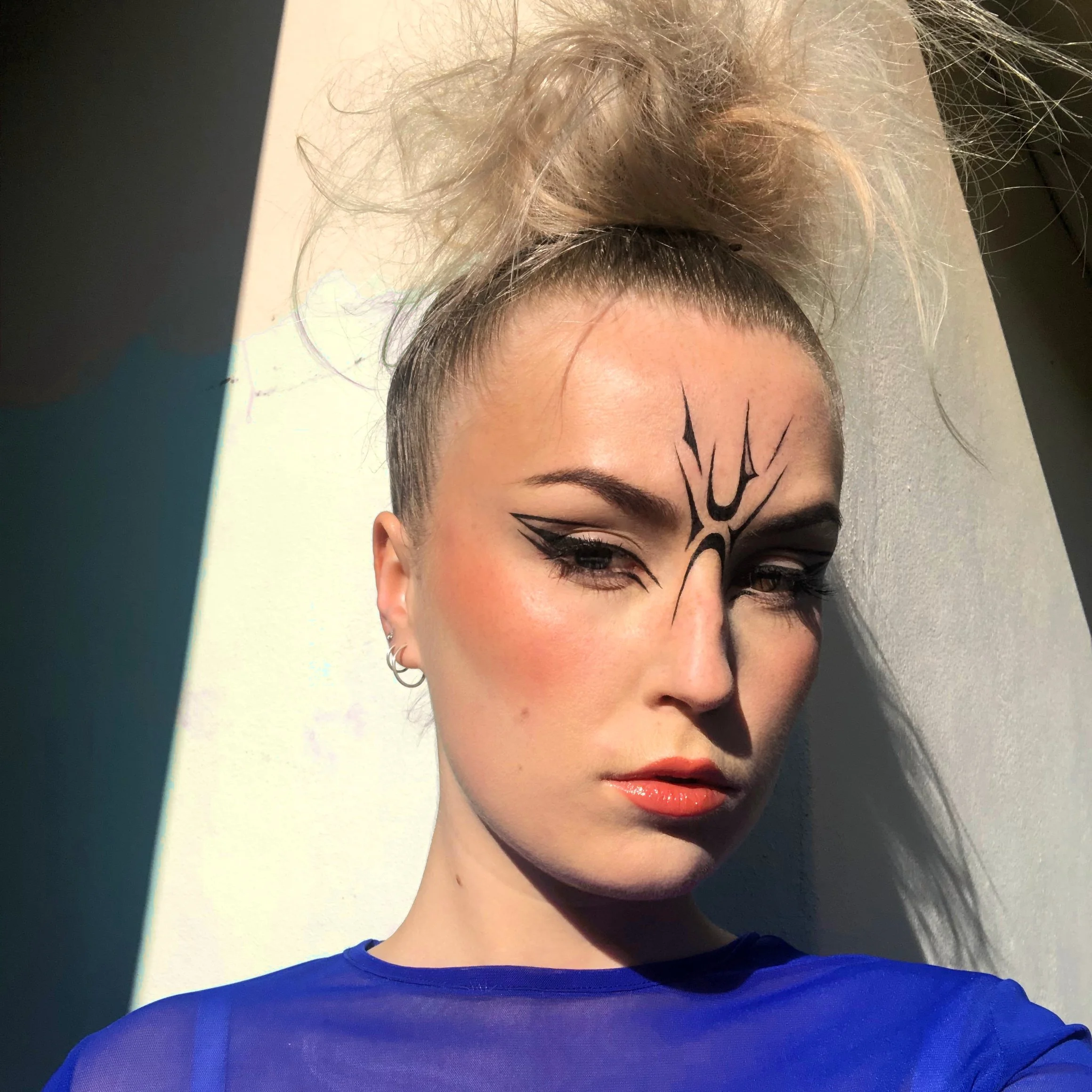 A woman with elaborate black face makeup and painted lines on her forehead and cheeks. She has light skin, blonde hair styled in a high messy bun, and is wearing a royal blue top. Sunlight is casting shadows on her face and background.