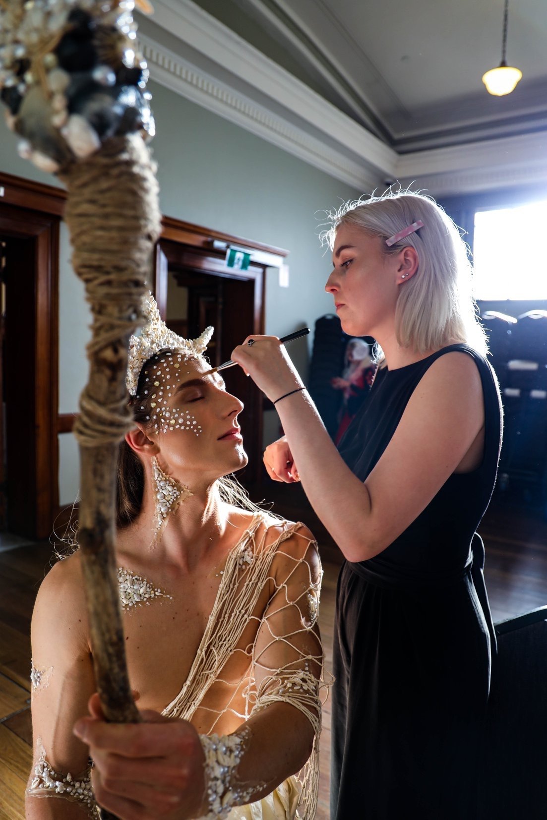 A woman with elaborate makeup and costume is getting her face painted or adorned by a makeup artist in a room with wooden floors and large windows.
