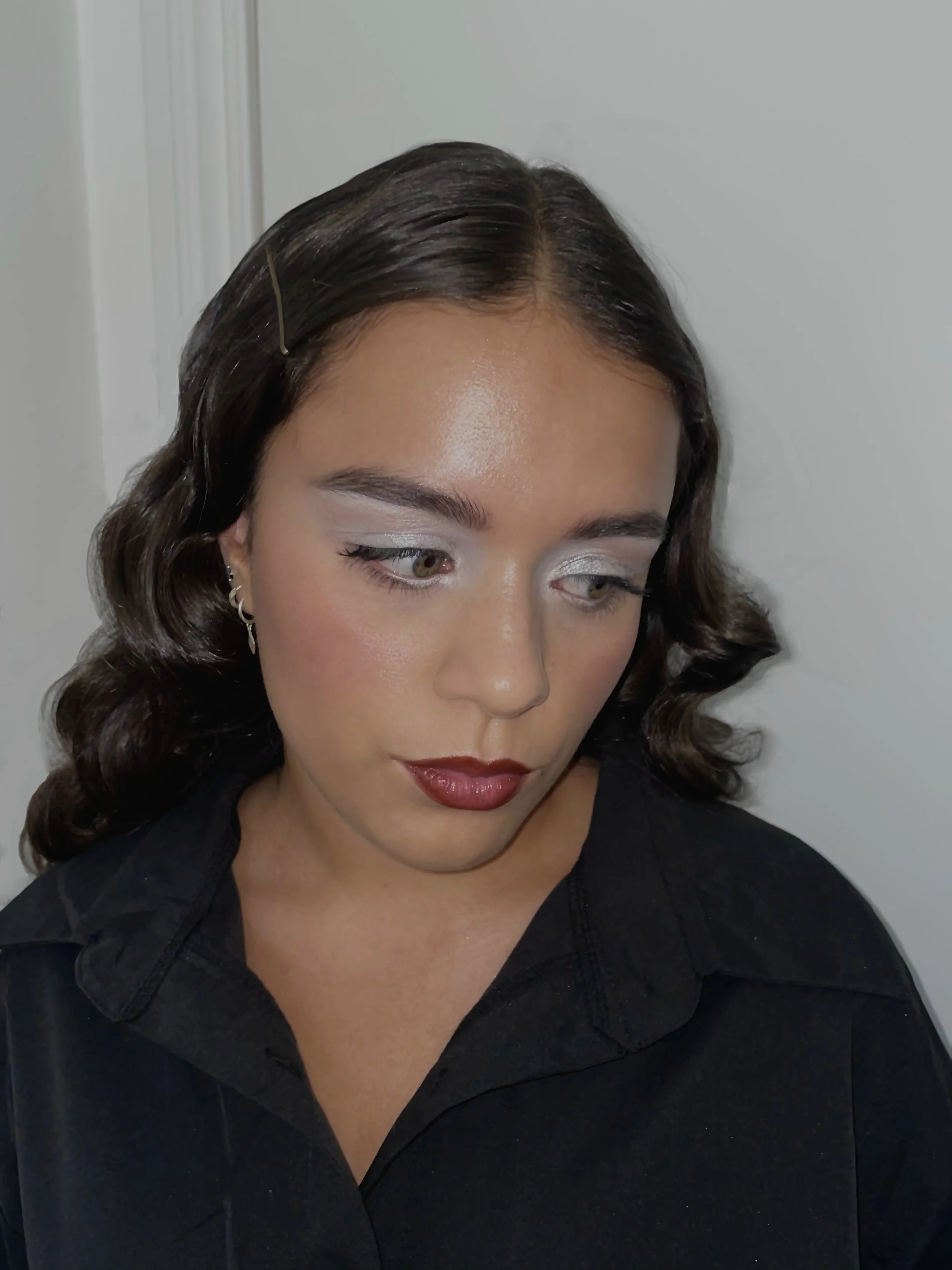 A woman with dark wavy hair, wearing makeup with silver eyeshadow and dark red lipstick, looking downward, dressed in a black shirt, standing against a plain background.