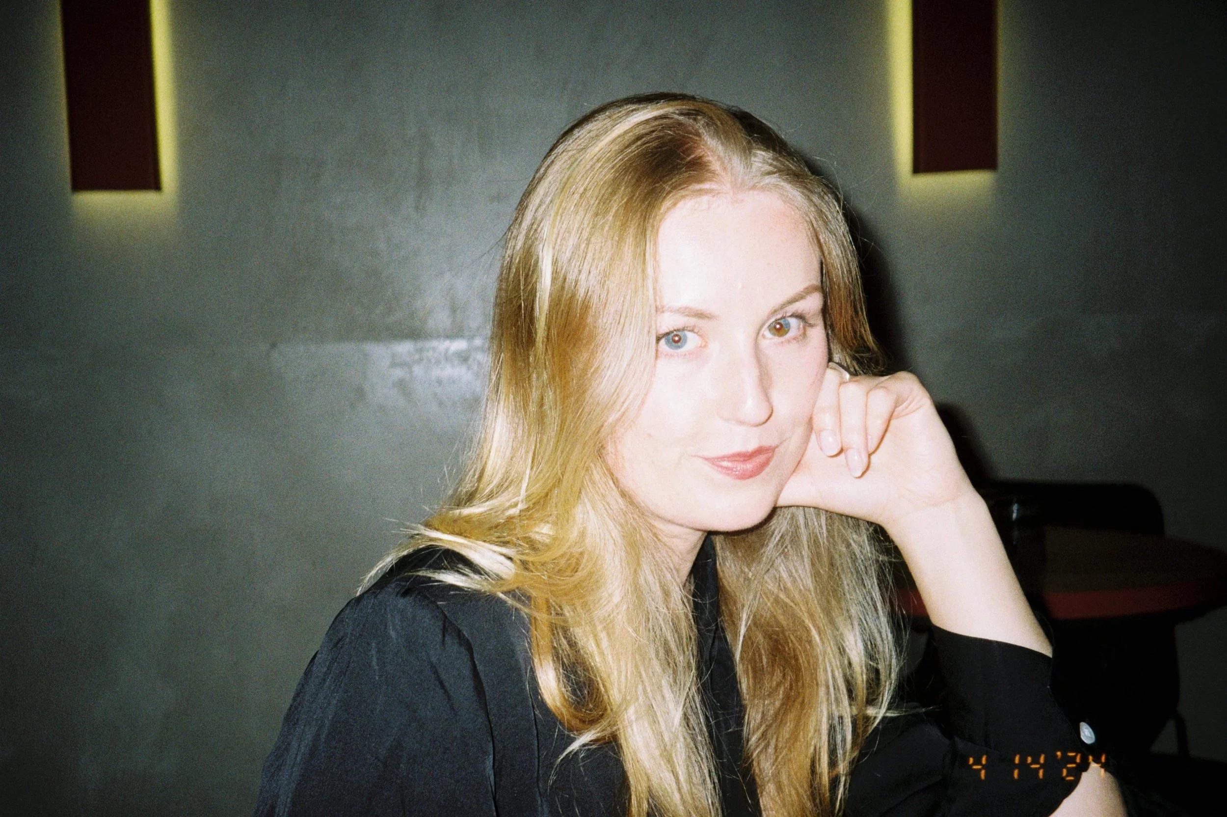 A woman with blonde hair, wearing a black top, resting her chin on her hand, smiling slightly, in a dimly lit room with dark walls.