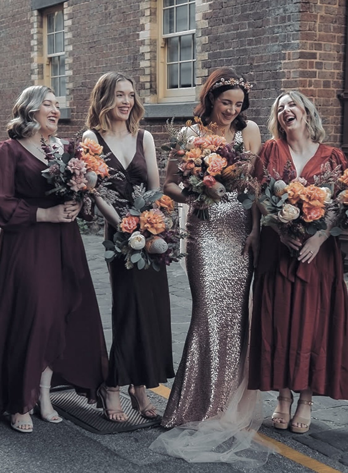 A group of five women at a wedding, with one woman in a sequin gown, holding bouquets, all smiling and laughing outside a brick building.