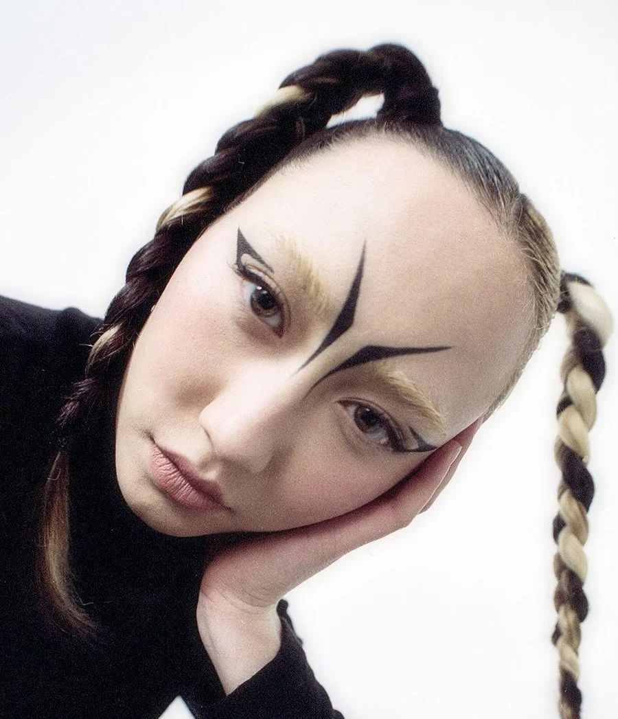 A young woman with elaborate geometric makeup on her face, dark and blonde braided hair, resting her head on her hand, against a plain background.
