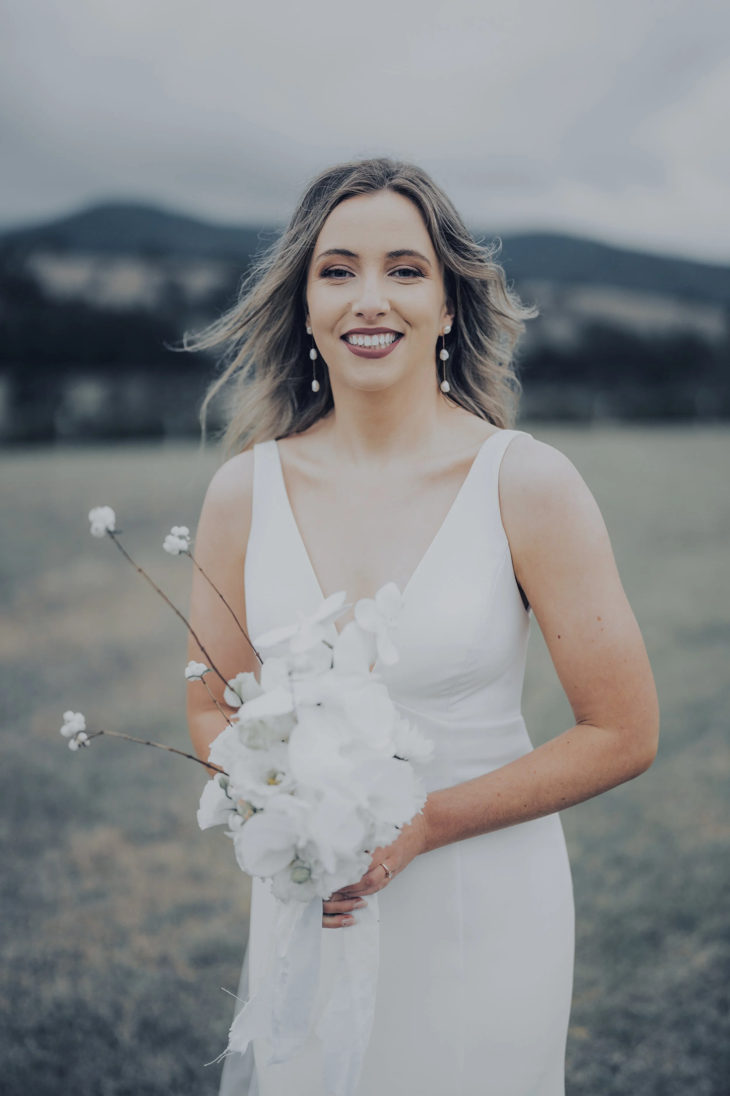 A woman in a white dress holding a bouquet of white flowers outdoors with a blurred landscape and hills in the background.